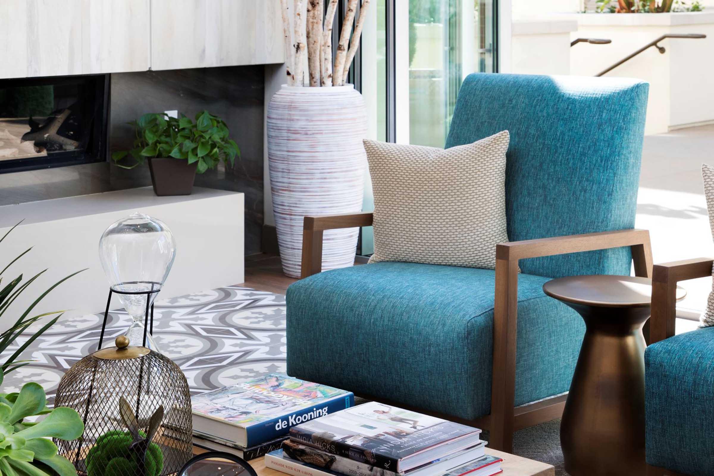 A cozy modern living space featuring a blue upholstered armchair with wooden arms, a decorative pillow, a brass side table, and a variety of books and magazines stacked on the floor. Green plants and a stylish rug add warmth to the room, complemented by a light fixture in the foreground.