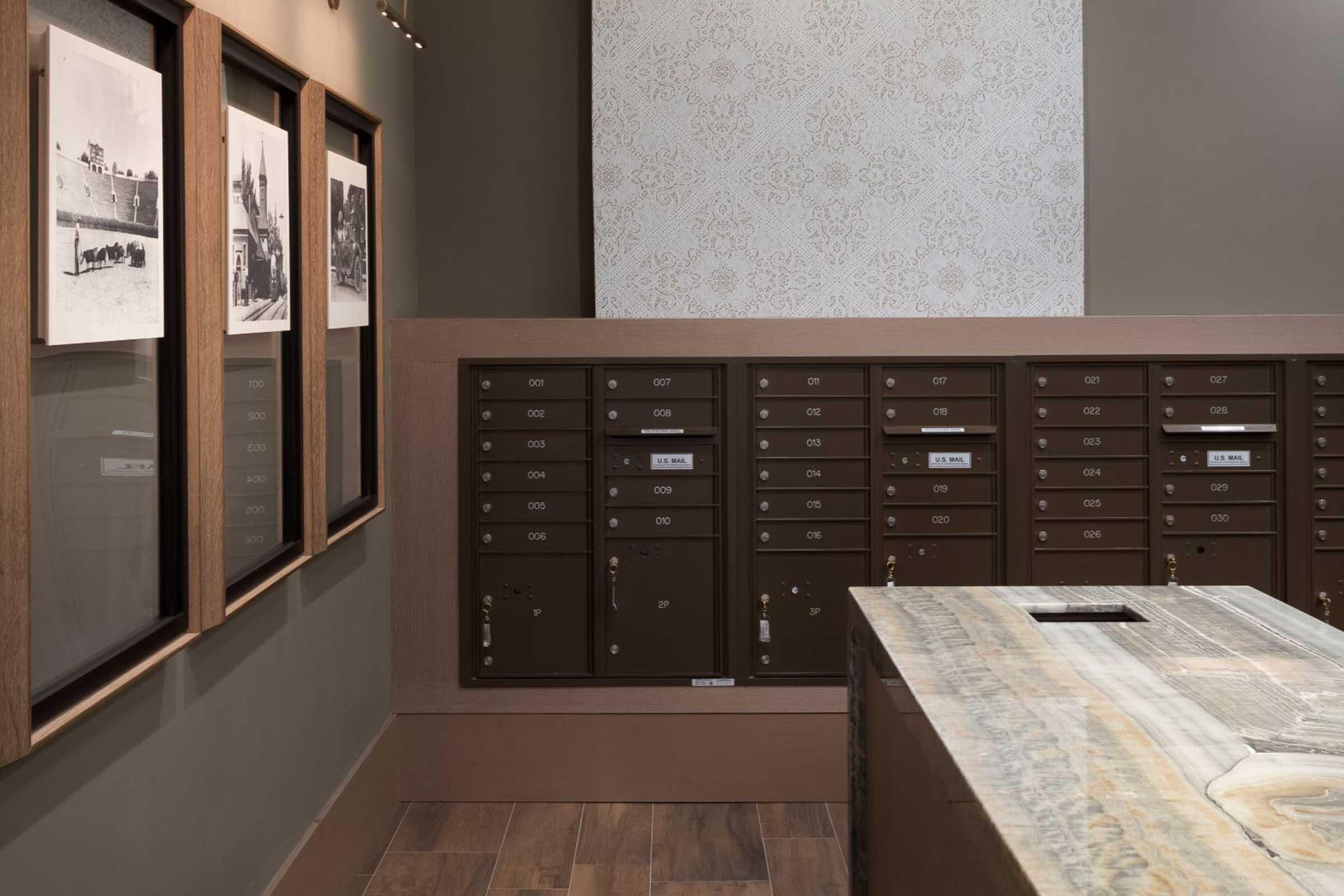 Interior view of a modern space featuring a wall of mailboxes. The wall is adorned with framed black-and-white photographs, and there is a wooden counter in the foreground. The decor combines contemporary and vintage elements, with muted colors and subtle design patterns on the wall behind the mailboxes.