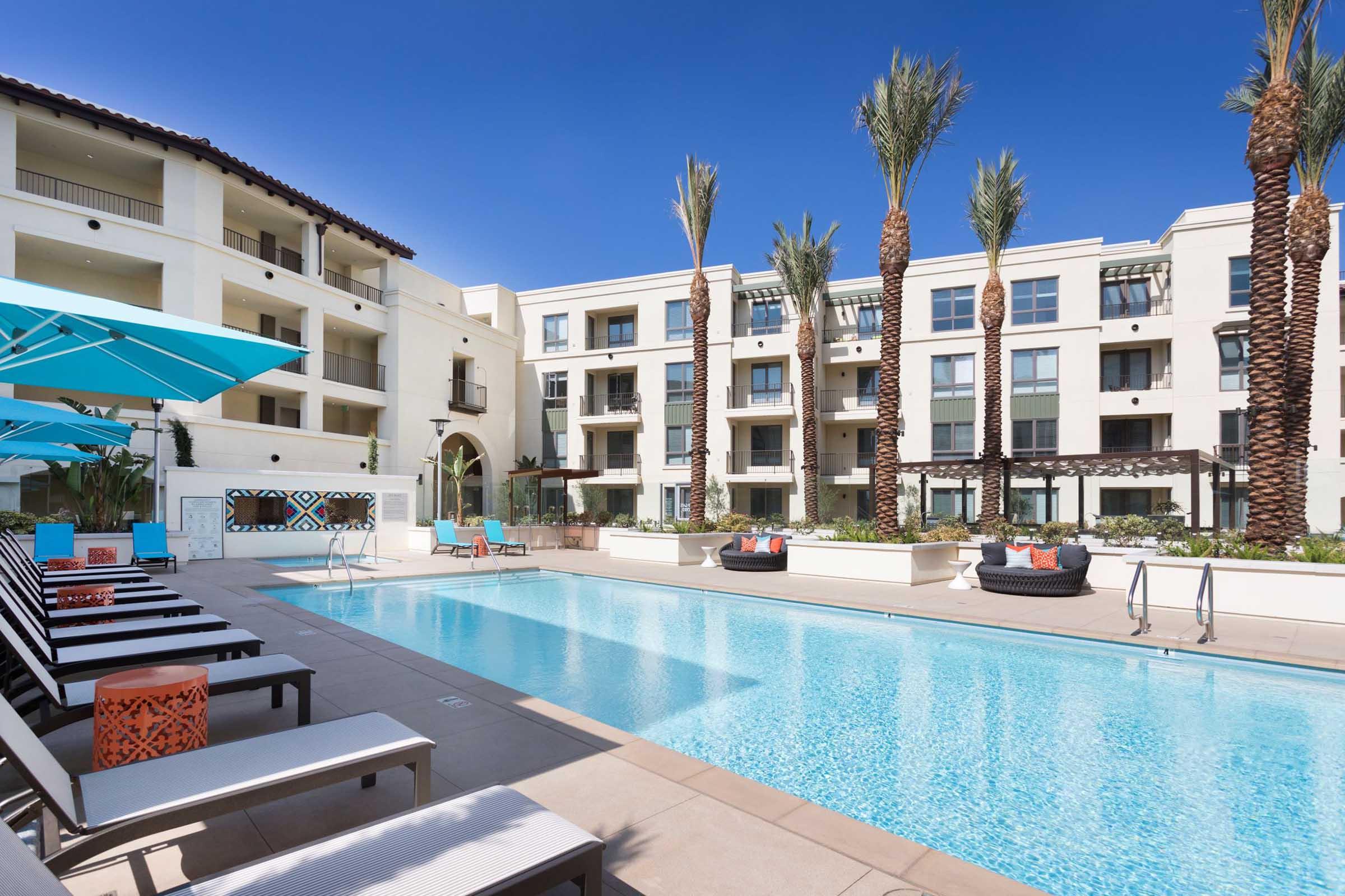 A bright, modern swimming pool surrounded by lounge chairs and umbrellas, with palm trees lining the property. The scene features a stylish apartment complex in the background, showcasing contemporary architecture and landscaping. Clear blue sky enhances the vibrant atmosphere.