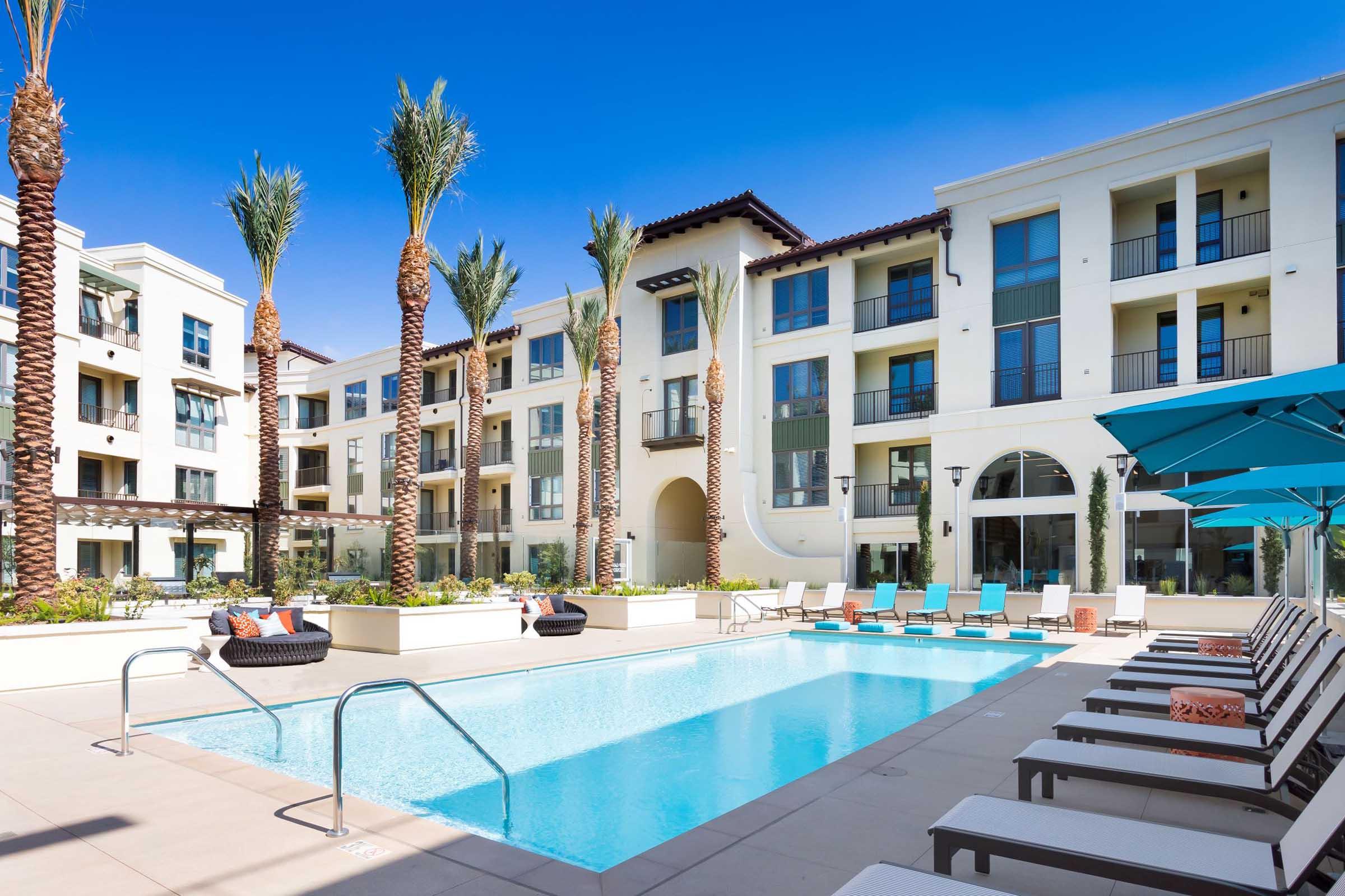 Modern apartment complex featuring a sparkling swimming pool surrounded by palm trees and lounge chairs. The buildings are designed in a contemporary style with multiple balconies, offering a bright and inviting atmosphere. Sun umbrellas provide shade near the pool area.