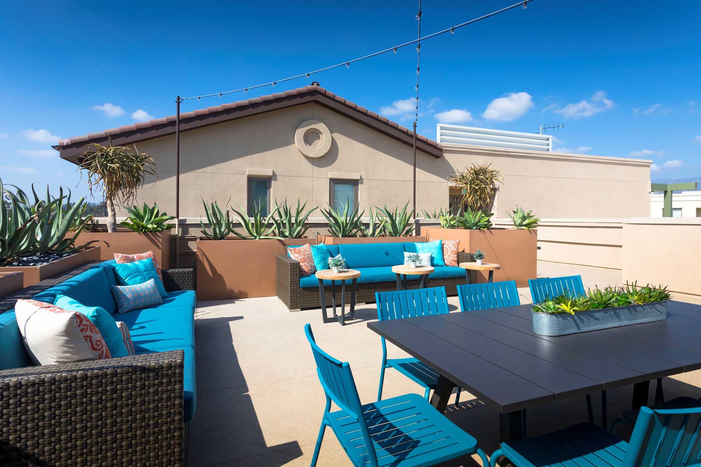 Rooftop terrace featuring comfortable seating with turquoise cushions, a dining table, and potted plants. The space is bright and inviting, with clear blue skies in the background and decorative string lights adding ambiance.