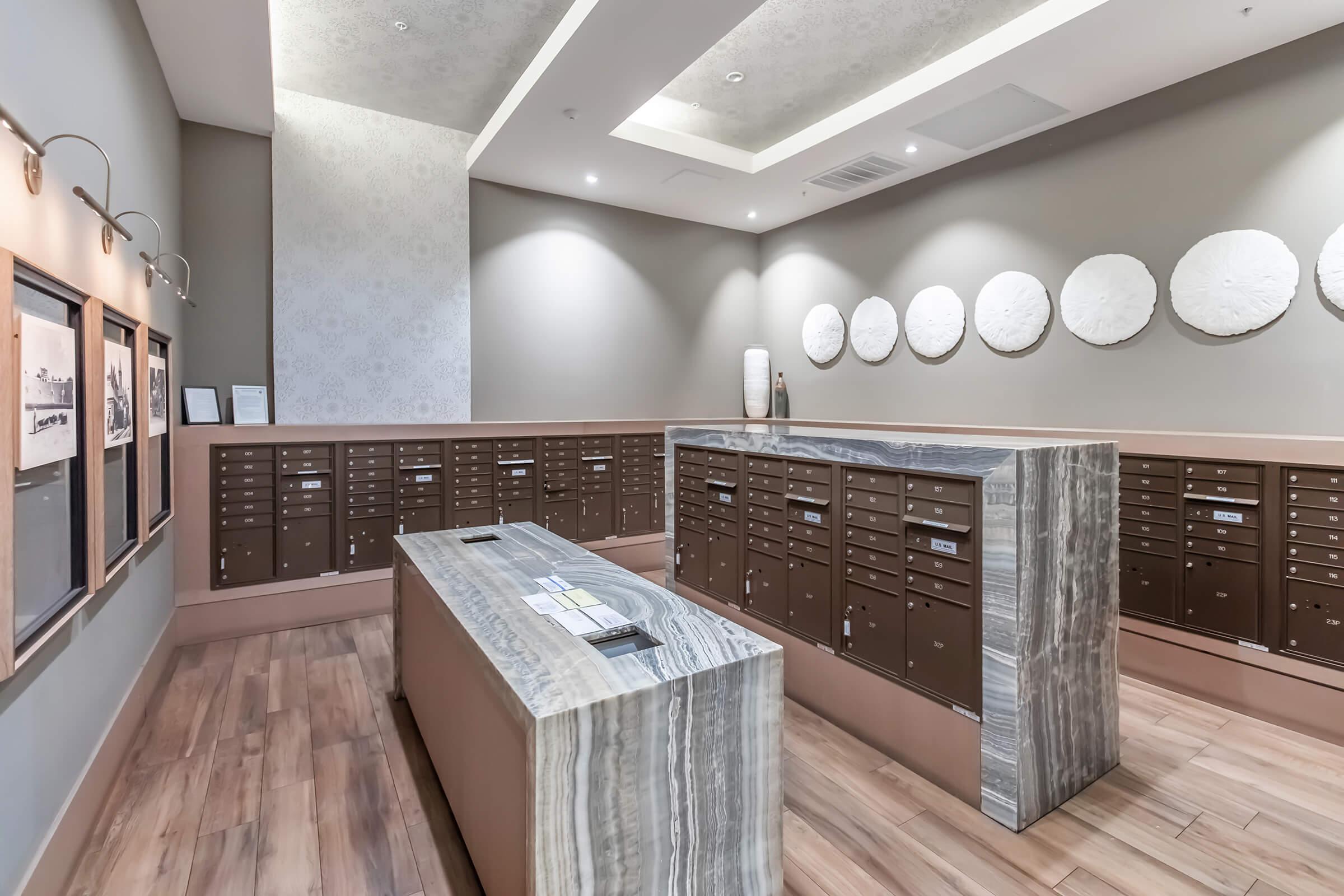 A modern mailroom featuring a marble counter and numerous secure mailboxes lined against the walls. The space is well-lit with soft gray tones, decorative wall art in circular shapes, and a wooden floor, creating a contemporary and organized atmosphere for residents to retrieve their mail.