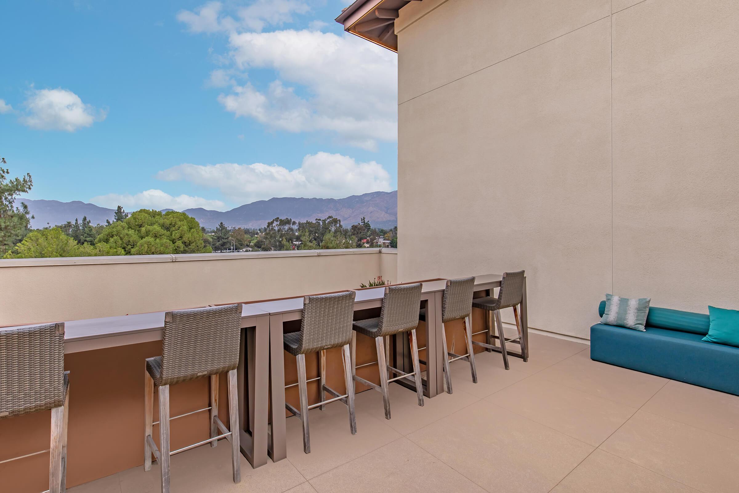 A spacious outdoor patio featuring a long bar with high stools and a cozy couch. The backdrop includes lush green trees and distant mountains under a partly cloudy sky, creating a tranquil setting for relaxation and socializing.