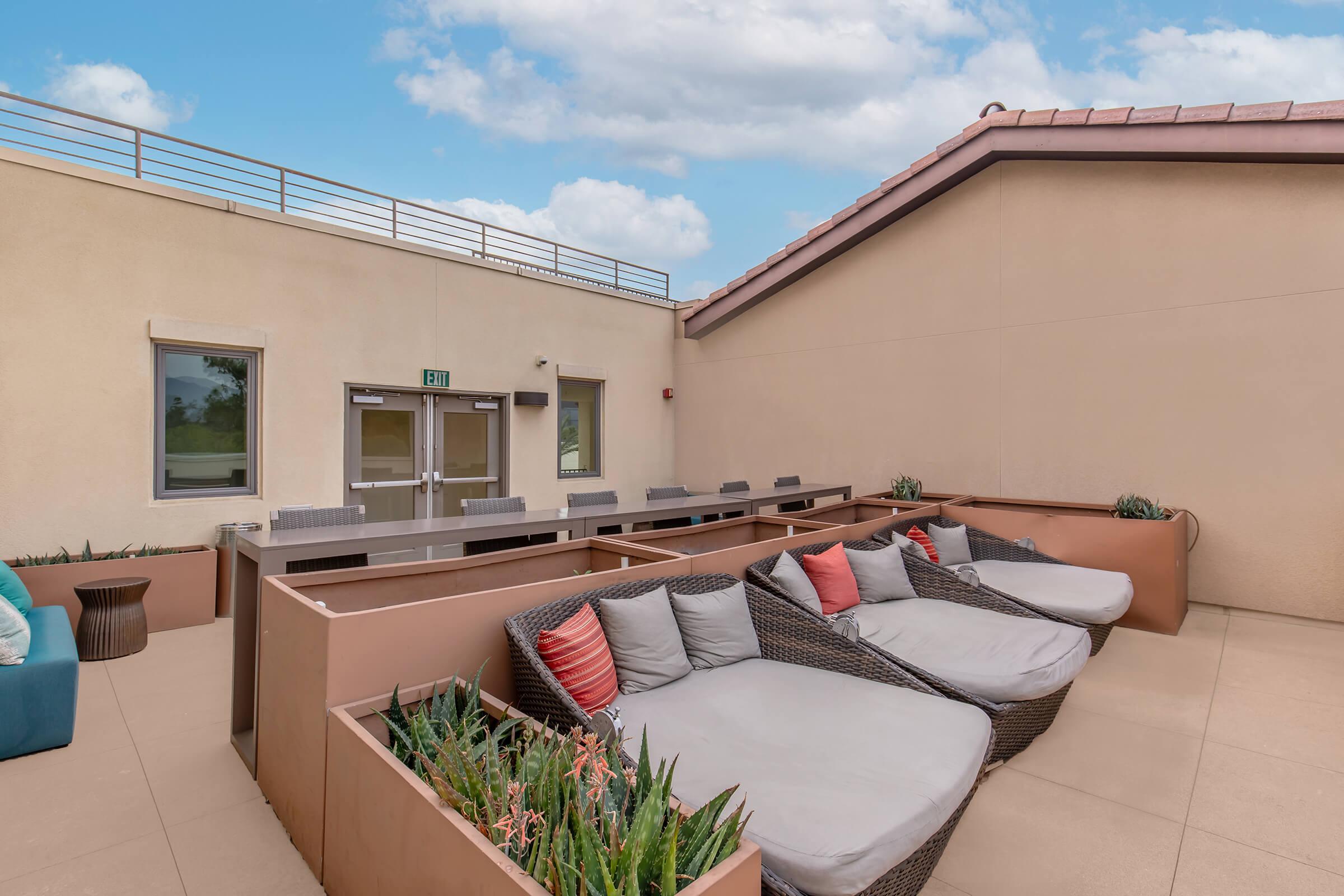 A stylish outdoor lounge area featuring large, comfortable seating with cushions, surrounded by planters with greenery. The space is well-lit and includes modern furniture, making it ideal for relaxation or social gatherings. A clear sky provides a bright backdrop.