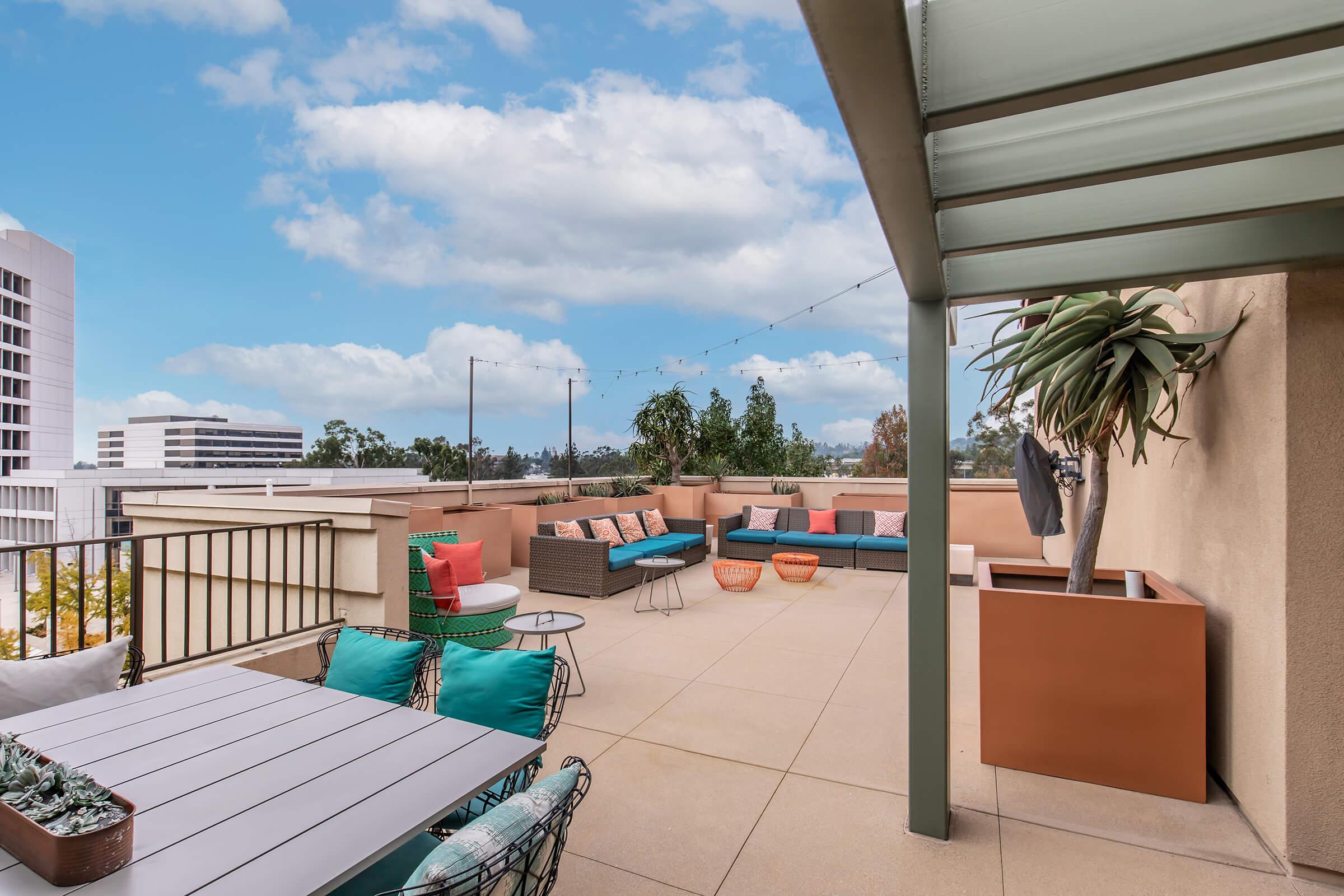 A rooftop patio with colorful outdoor furniture, including a seating area with cushions, a dining table, and decorative potted plants. The backdrop features a clear blue sky and distant buildings, creating a relaxed and inviting atmosphere.