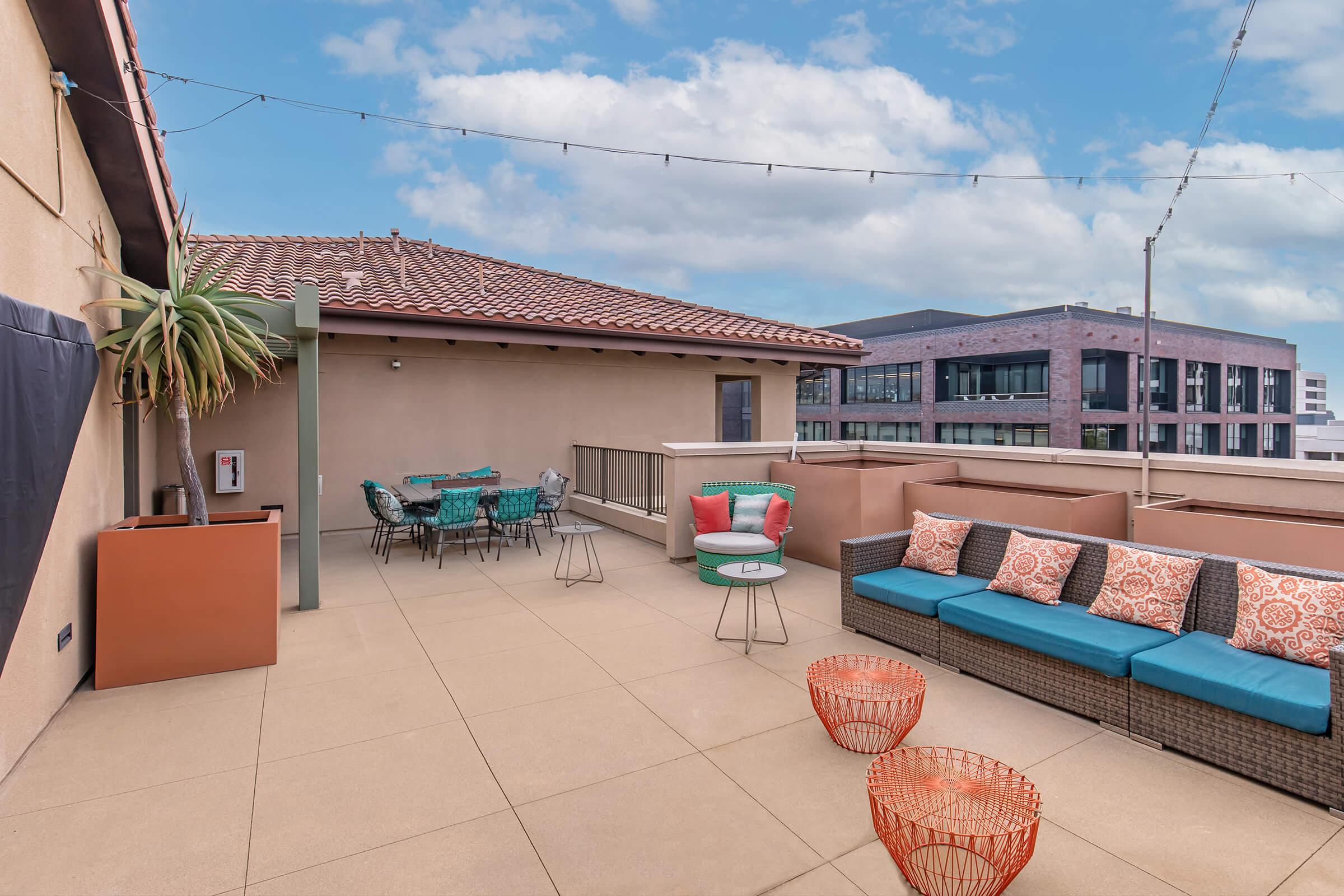 A spacious rooftop terrace featuring modern outdoor furniture, including a large sectional sofa with colorful cushions, stylish chairs, and tables. Decorative elements include potted plants and string lights overhead, set against a backdrop of a blue sky and contemporary buildings.