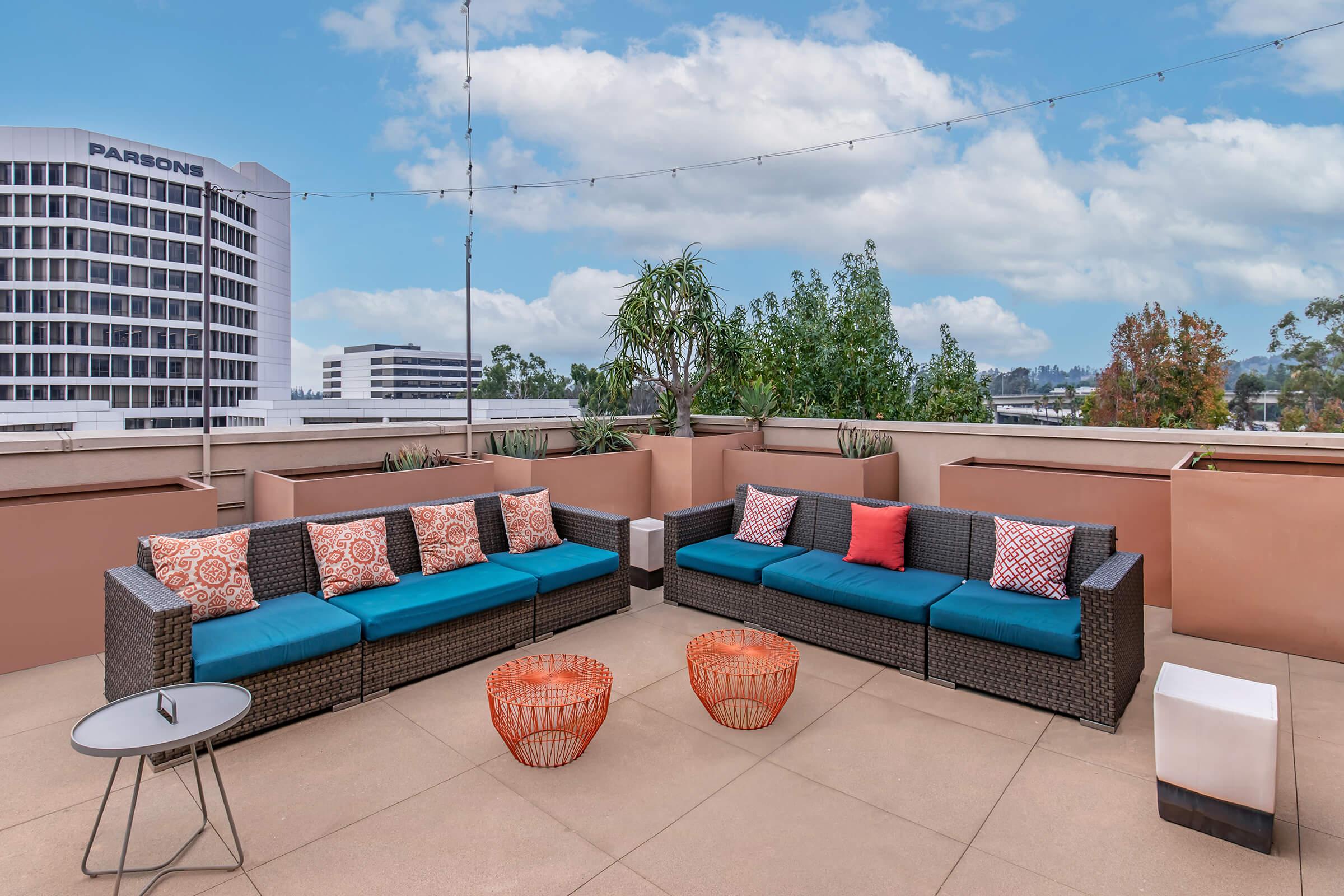 A rooftop patio featuring two large wicker seating areas with blue cushions and colorful pillows, surrounded by potted plants. There are two orange wire coffee tables and a small round table. The background shows a building labeled "PARSONS" and a clear blue sky with scattered clouds.