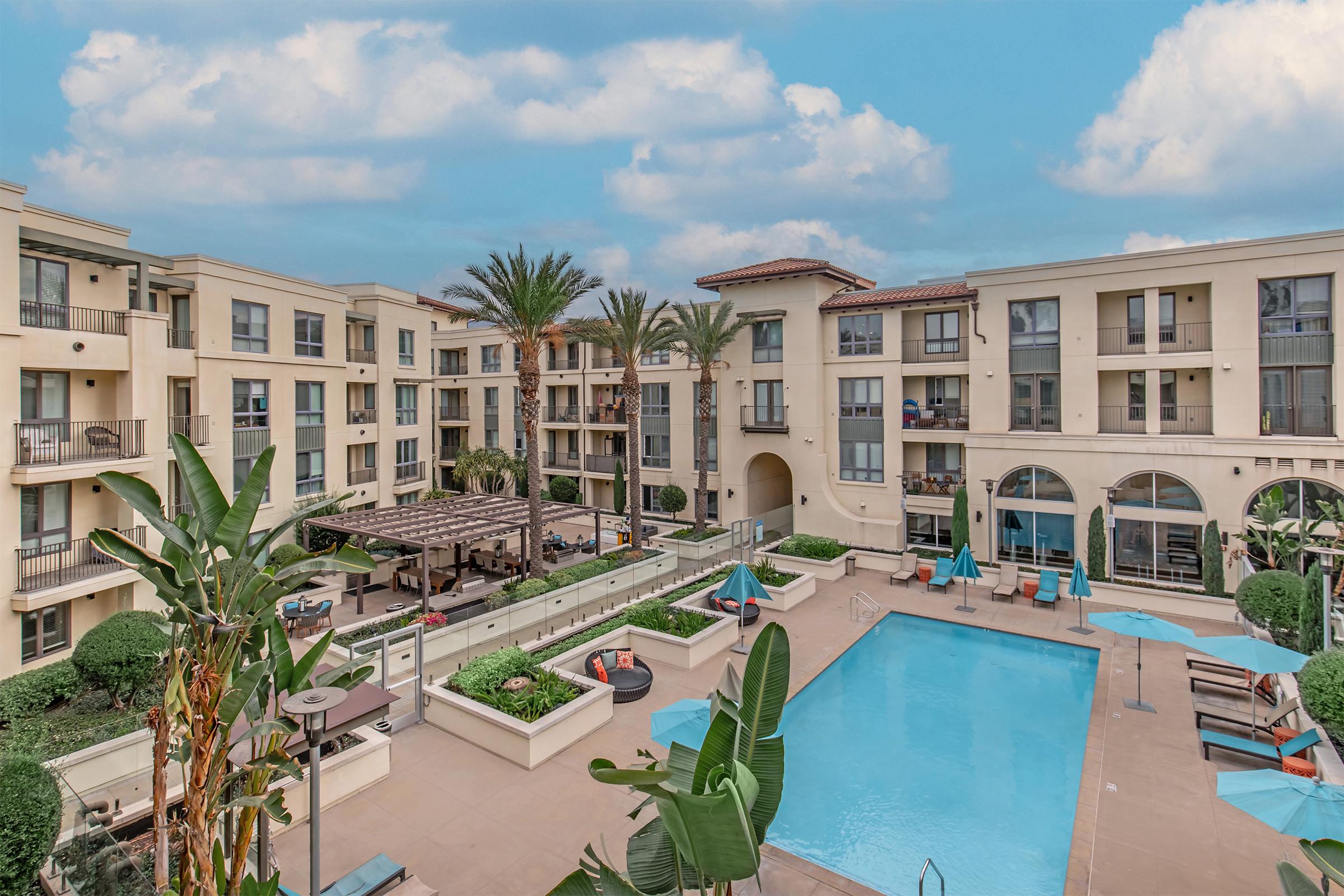 Aerial view of a modern apartment complex featuring a swimming pool surrounded by lounge chairs and palm trees. Balconies are visible on the buildings, which have a light-colored facade and arched windows. The sky is partly cloudy, adding a vibrant atmosphere to the scene.