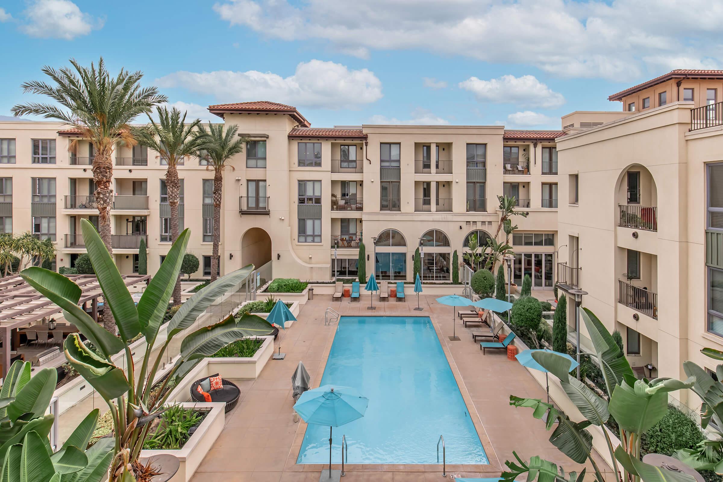 A view of a luxurious courtyard featuring a swimming pool surrounded by palm trees and lush greenery. The area includes lounge chairs, umbrellas, and well-maintained landscaping, with modern apartment buildings in the background under a partly cloudy sky.