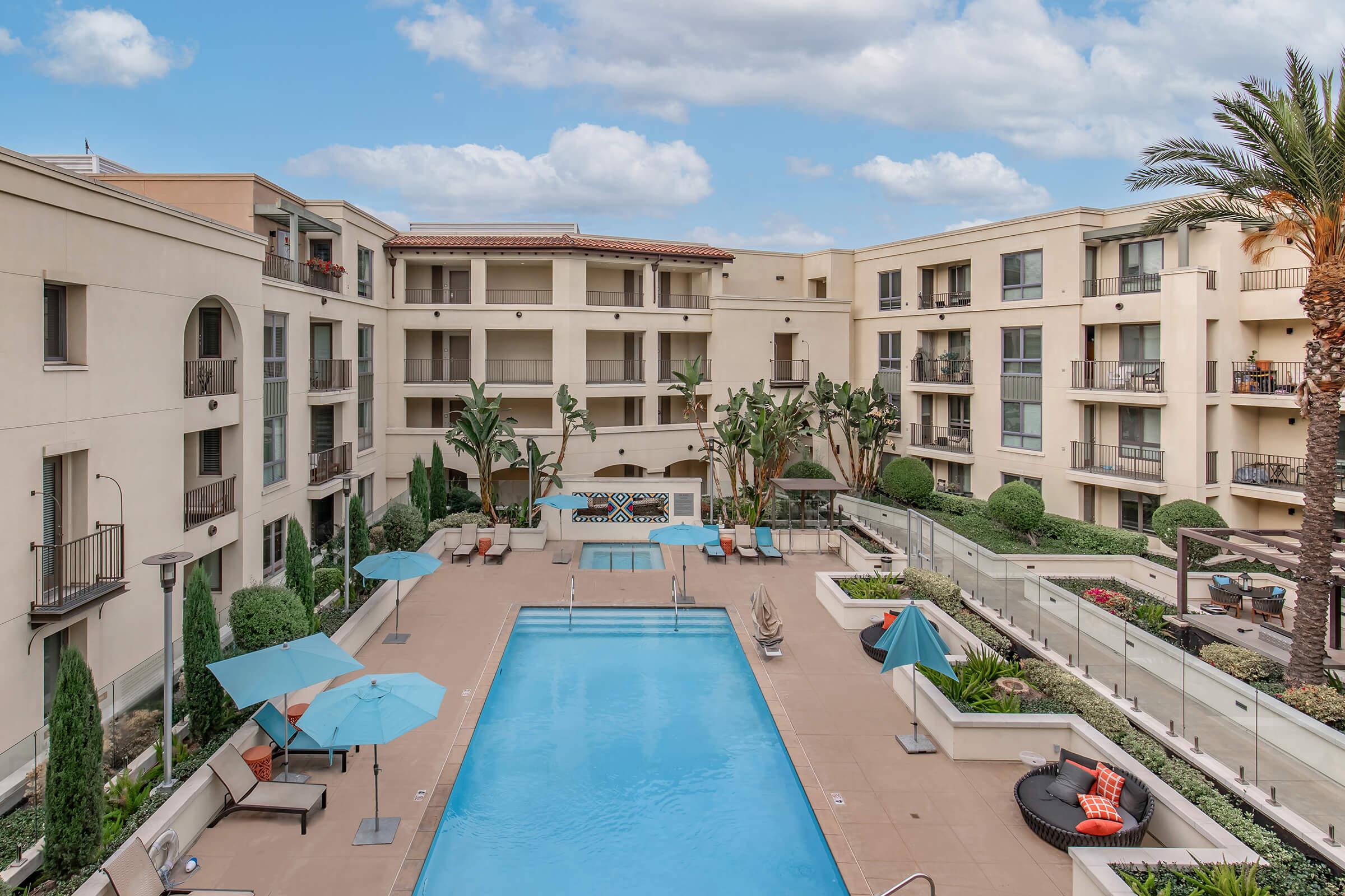 Aerial view of a modern apartment complex featuring a central swimming pool, palm trees, and lounge chairs. Umbrellas provide shade around the pool area, surrounded by landscaped gardens and buildings with balconies. The sky is partly cloudy, creating a bright and inviting atmosphere.