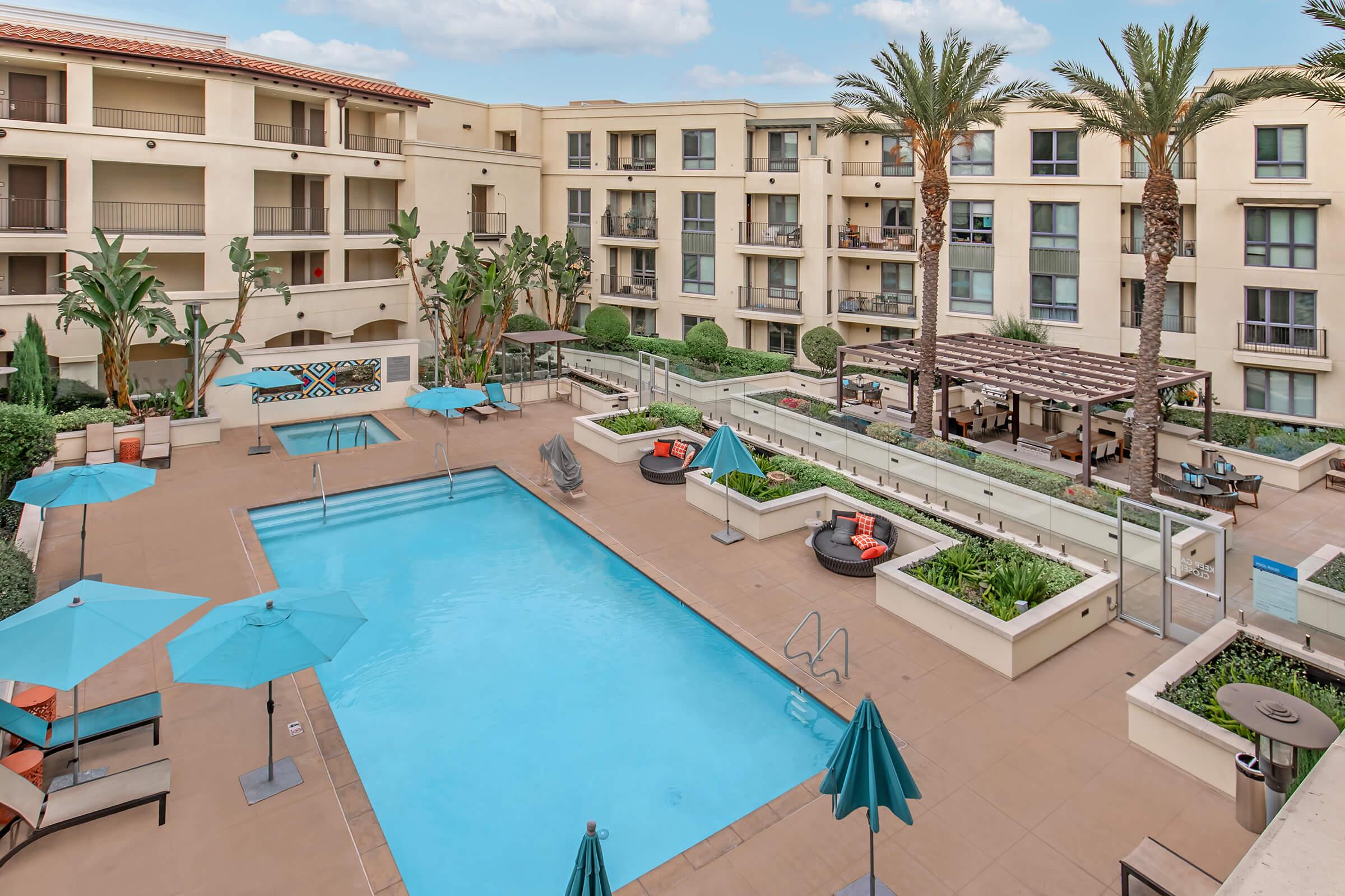 A spacious outdoor area featuring a swimming pool surrounded by lounge chairs and umbrellas. Lush greenery and palm trees are visible, along with a pergola and seating areas. The backdrop consists of a multi-story building with balconies and large windows.
