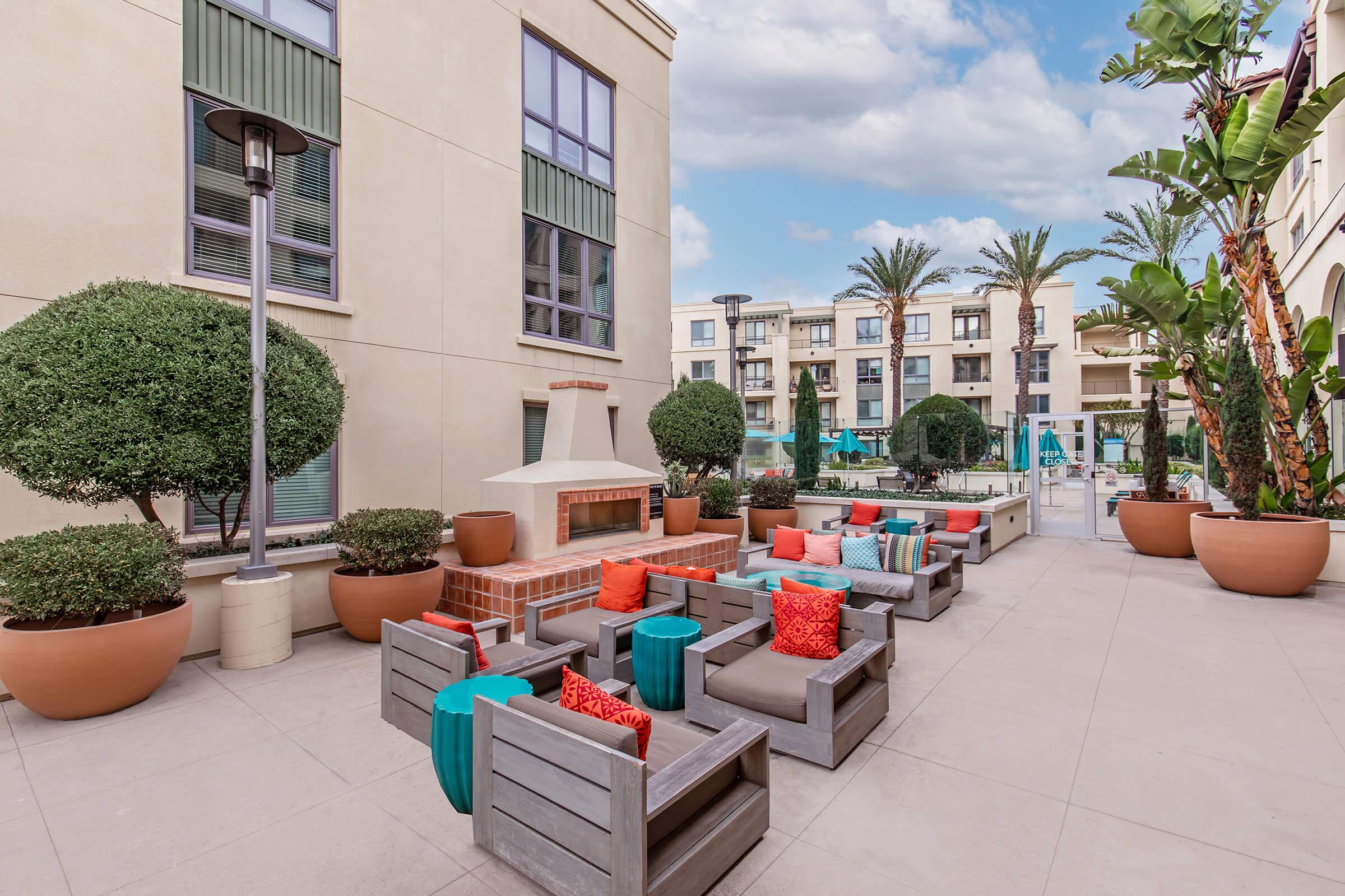 An outdoor lounge area featuring comfortable seating with colorful cushions, a fire pit, and neatly trimmed plants in large pots. The setting is bright and inviting, surrounded by palm trees and a swimming pool in the background, ideal for relaxation and social gatherings.