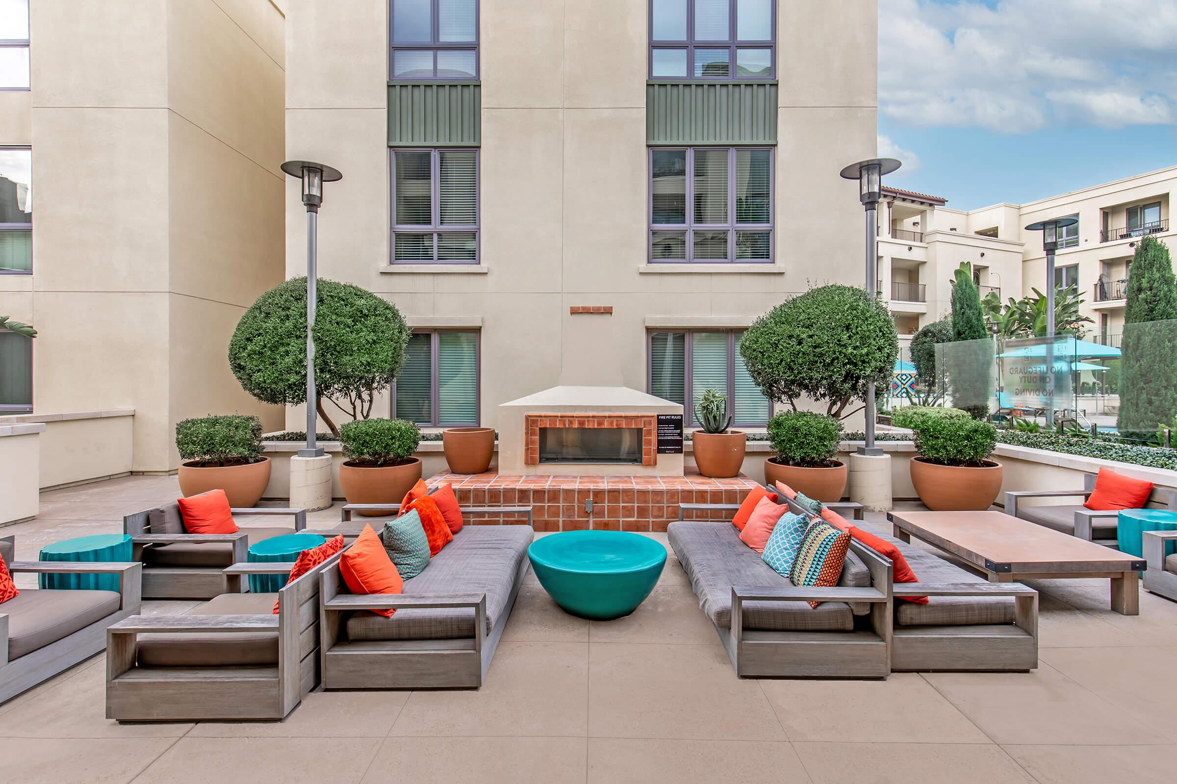 A stylish outdoor lounge area featuring modern seating with colorful cushions, a round turquoise table, and well-maintained greenery in large pots. A cozy fireplace is set against a backdrop of contemporary buildings, with tall lamps providing light. The space is inviting for relaxation and social gatherings.