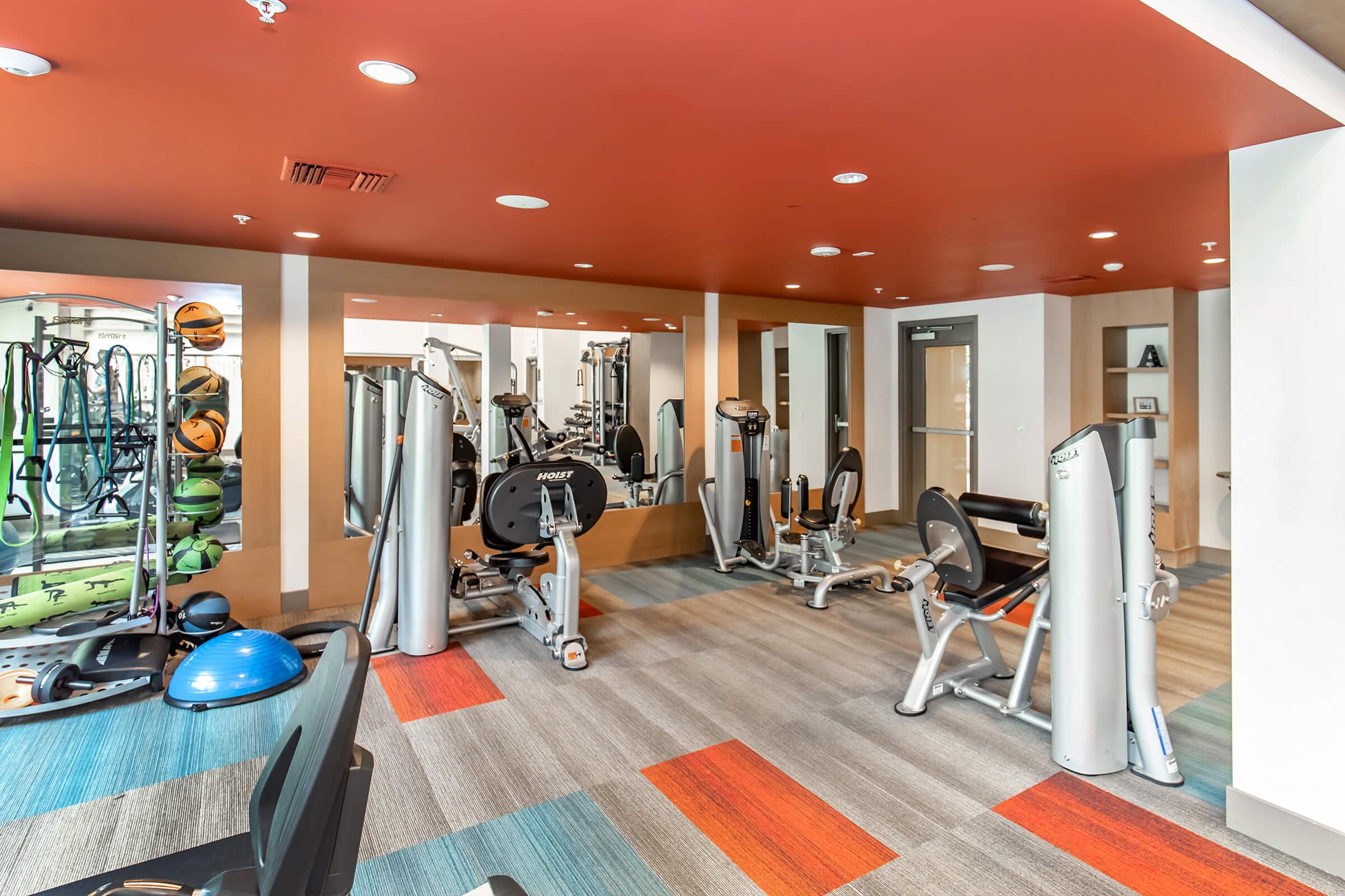 A modern gym interior featuring exercise machines, free weights, and fitness equipment. The space has bright orange and blue accents, mirrors, and a variety of gym accessories, creating an inviting environment for workouts.