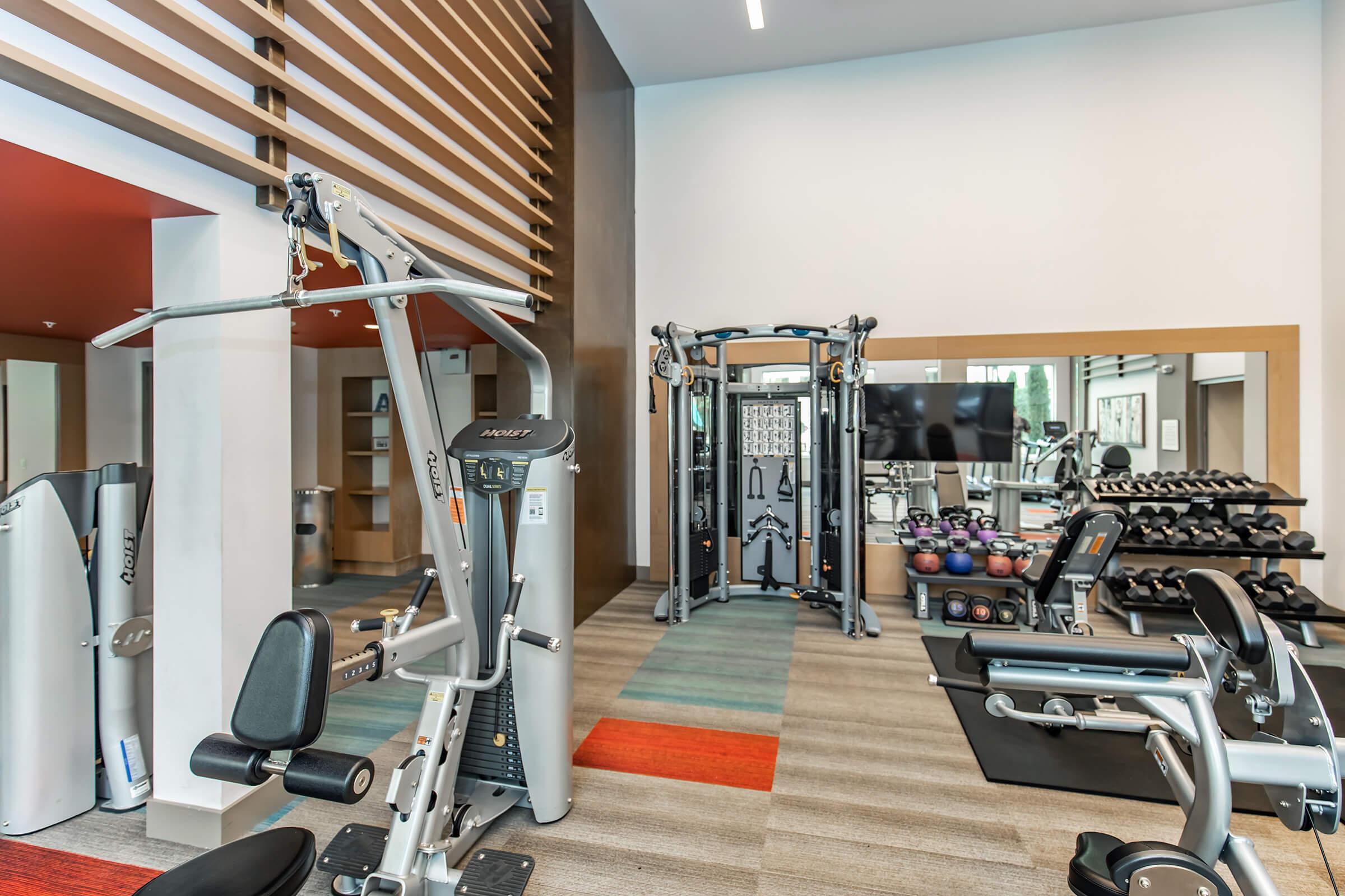 A modern gym interior featuring various workout equipment, including weight machines, dumbbells, a cable machine, and a mirror. The design includes a mix of light and dark wood accents with colorful flooring, creating a bright and inviting space for exercise.