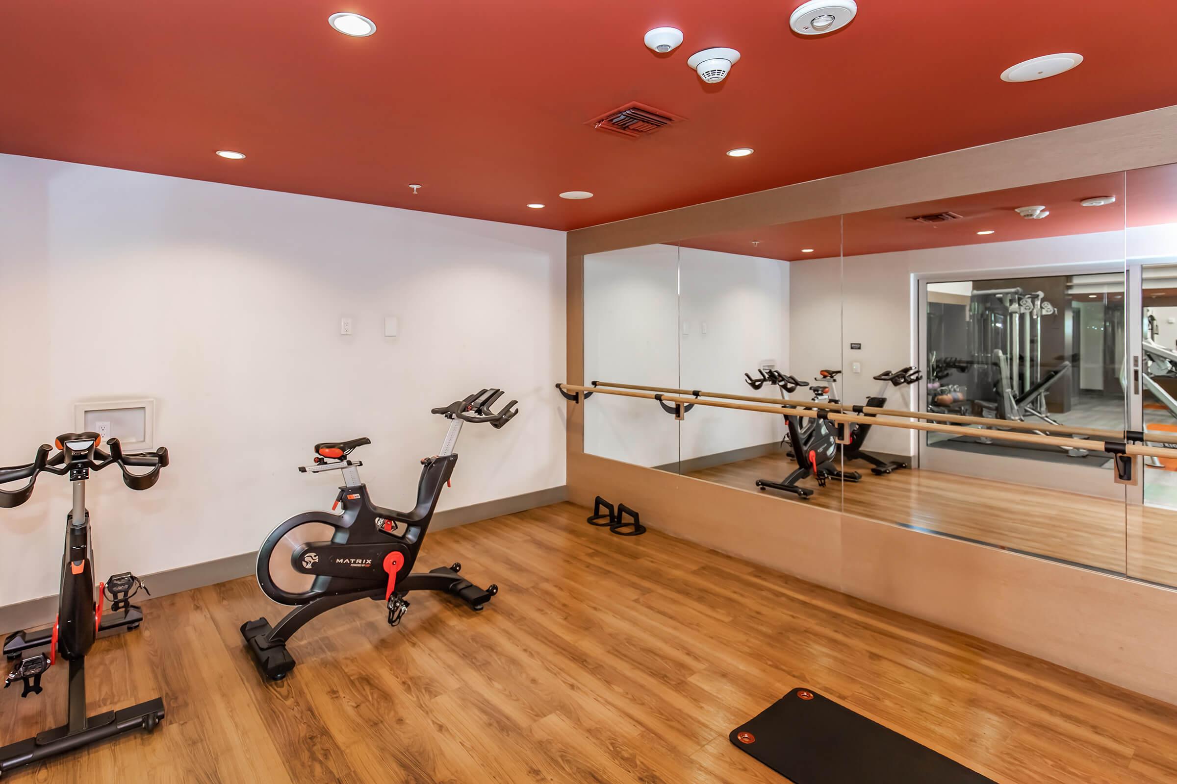 A modern fitness room featuring a stationary exercise bike, a mirror, and a wooden floor. Wall-mounted bars are visible, and the room is well-lit with a red ceiling. The setup suggests a space for workout classes or individual training.