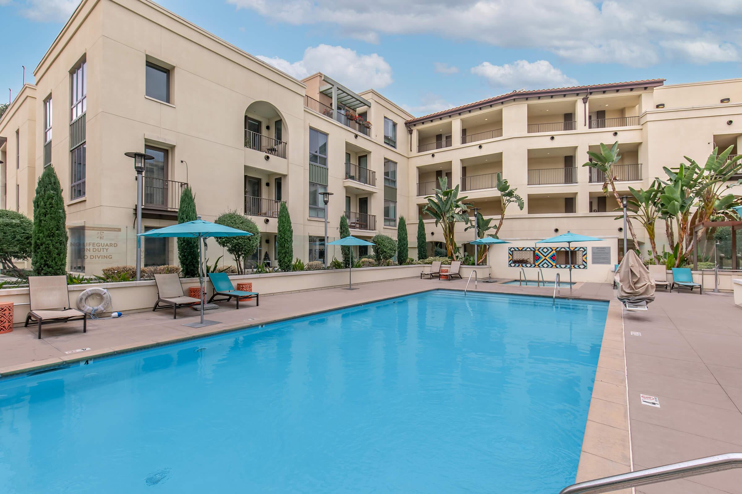 Swimming pool surrounded by modern apartment buildings, with lounge chairs and umbrellas. Lush greenery and palm trees provide a tropical feel. The scene is bright, showcasing a clear blue sky and well-maintained outdoor spaces designed for relaxation and leisure.
