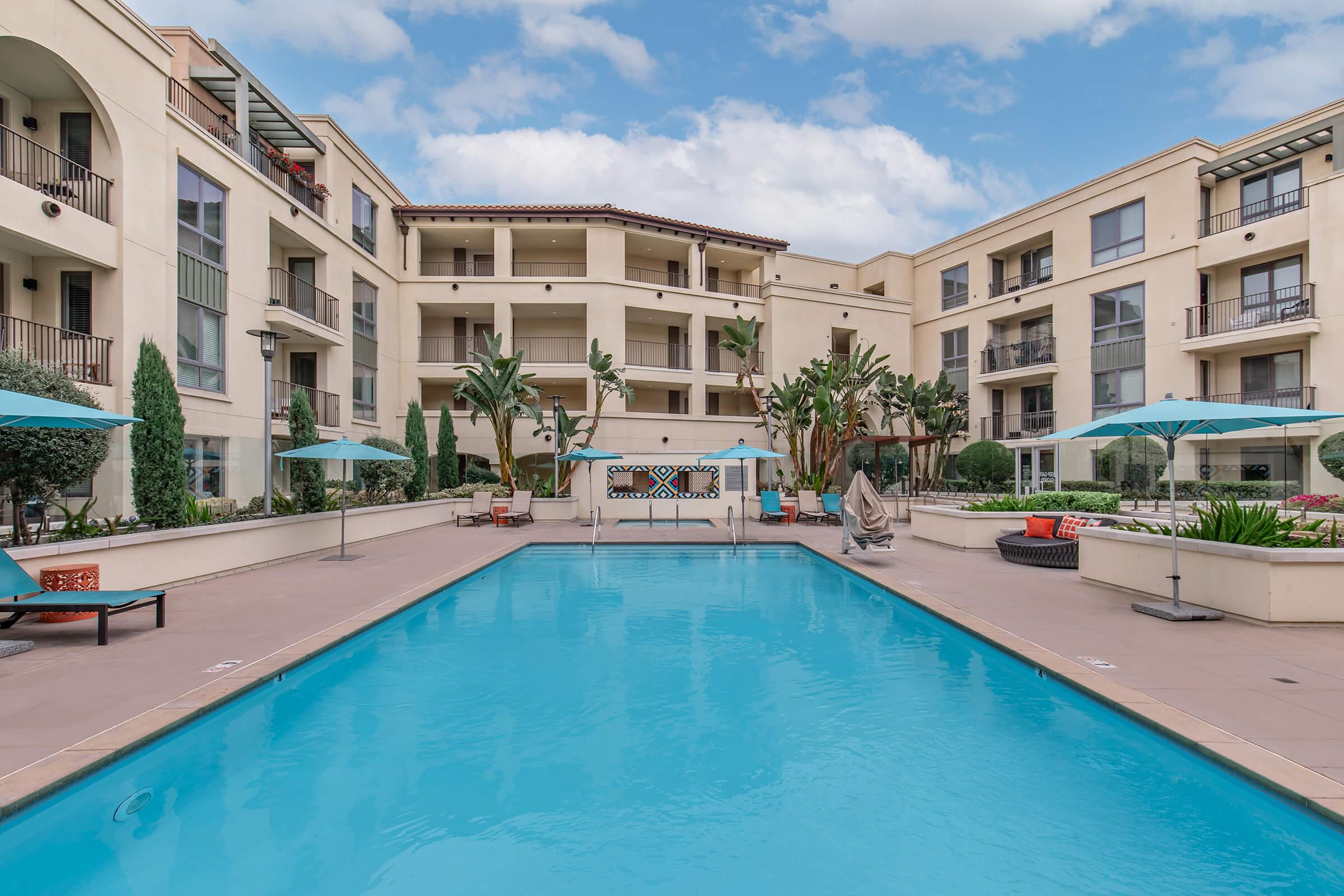 A serene courtyard featuring a large swimming pool surrounded by palm trees and modern apartment buildings. Lounge chairs and umbrellas are positioned near the pool, creating a relaxing outdoor space. The sky is partly cloudy, adding to the inviting atmosphere of the area.