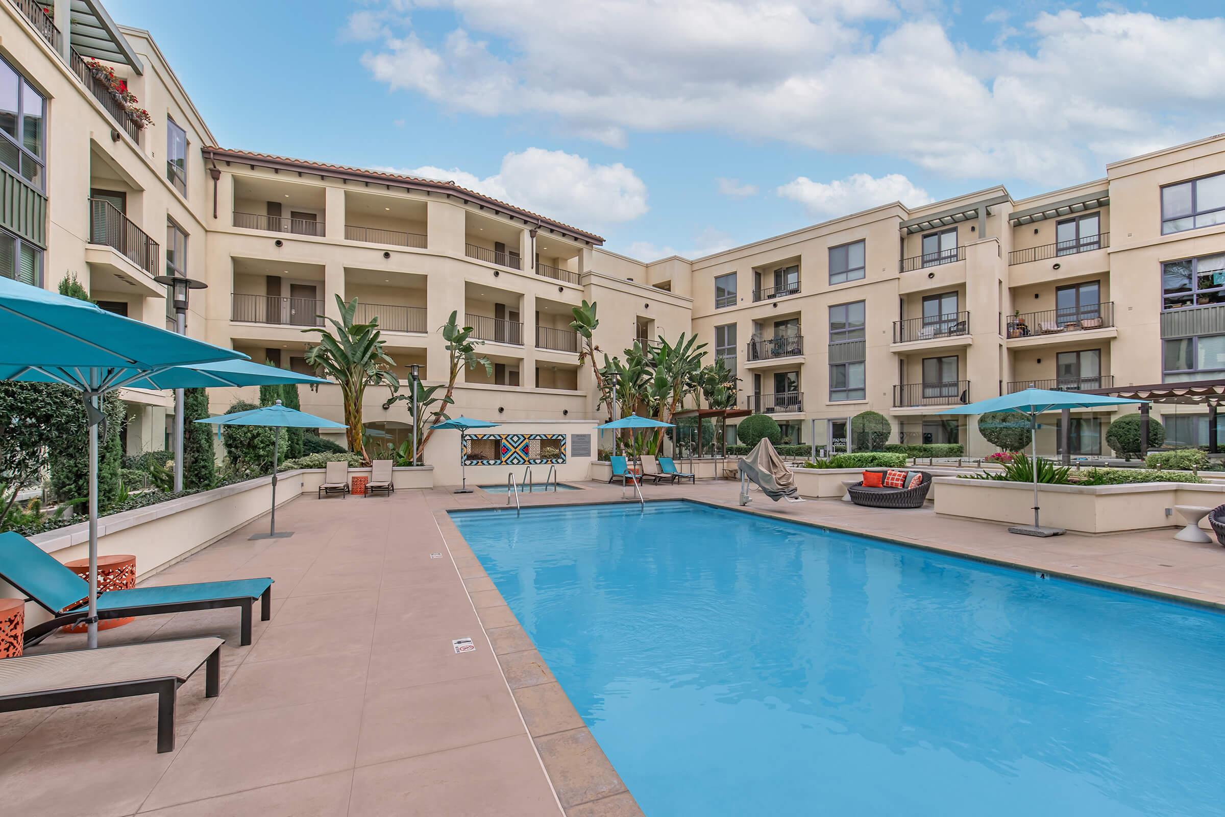 A view of a resort-style swimming pool surrounded by lounge chairs and umbrellas, with modern apartment buildings in the background. The area features lush greenery and decorative elements, creating a relaxing atmosphere in a sunny environment.
