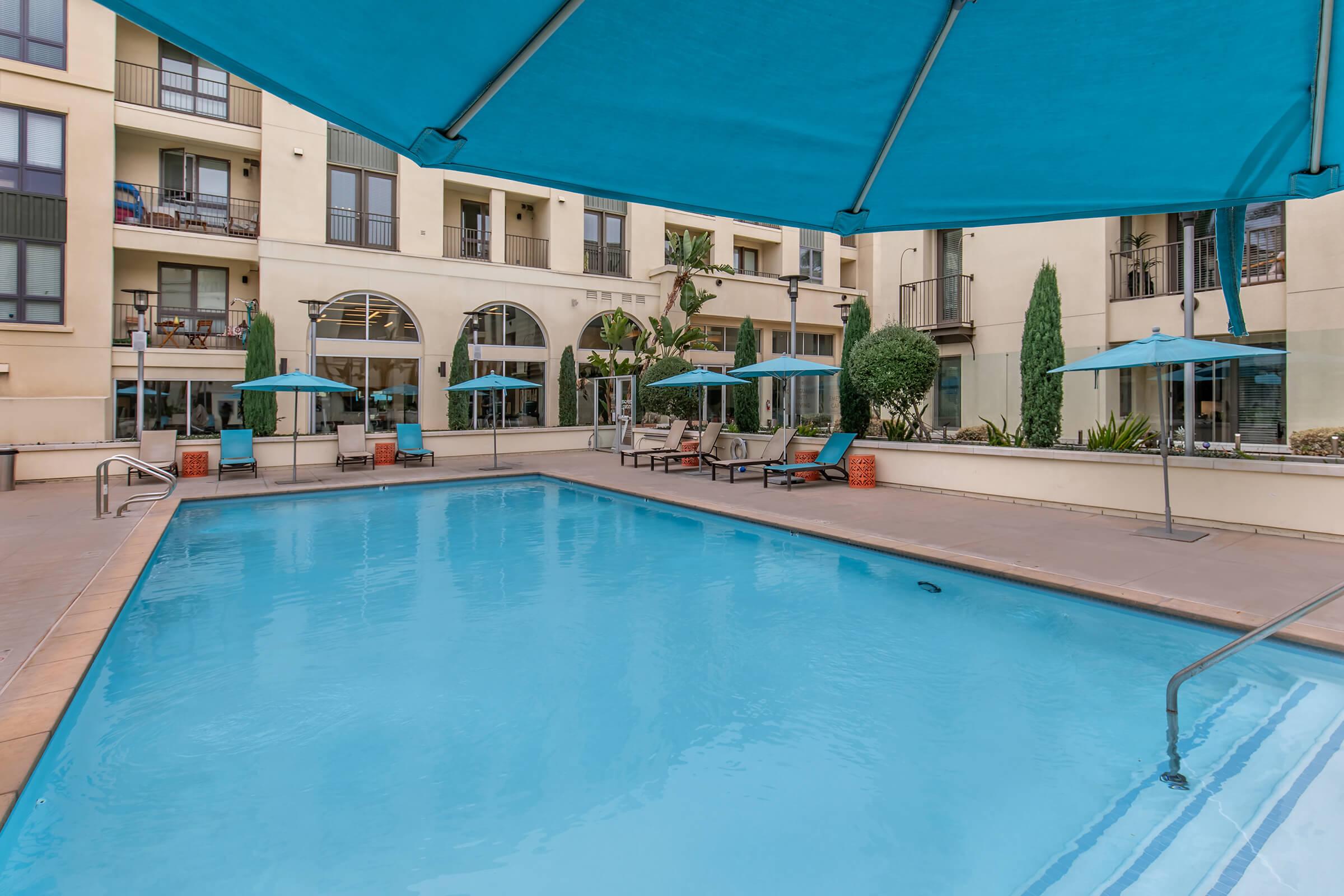 A swimming pool surrounded by lounge chairs and umbrellas, with a backdrop of a modern apartment building. The pool area features clear blue water and landscaped greenery, creating a relaxing outdoor atmosphere.