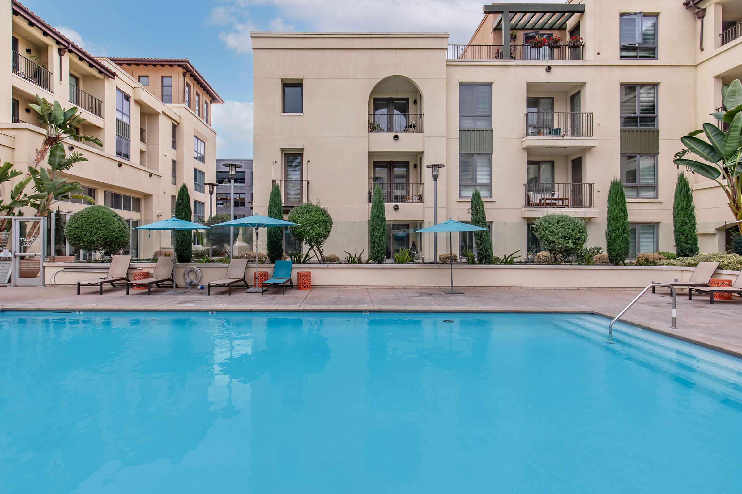 A clear blue swimming pool surrounded by lounge chairs and umbrellas, with modern apartment buildings in the background. Lush greenery includes small trees and shrubs, creating a serene outdoor space. The setting is bright and inviting, suitable for relaxation and socializing.