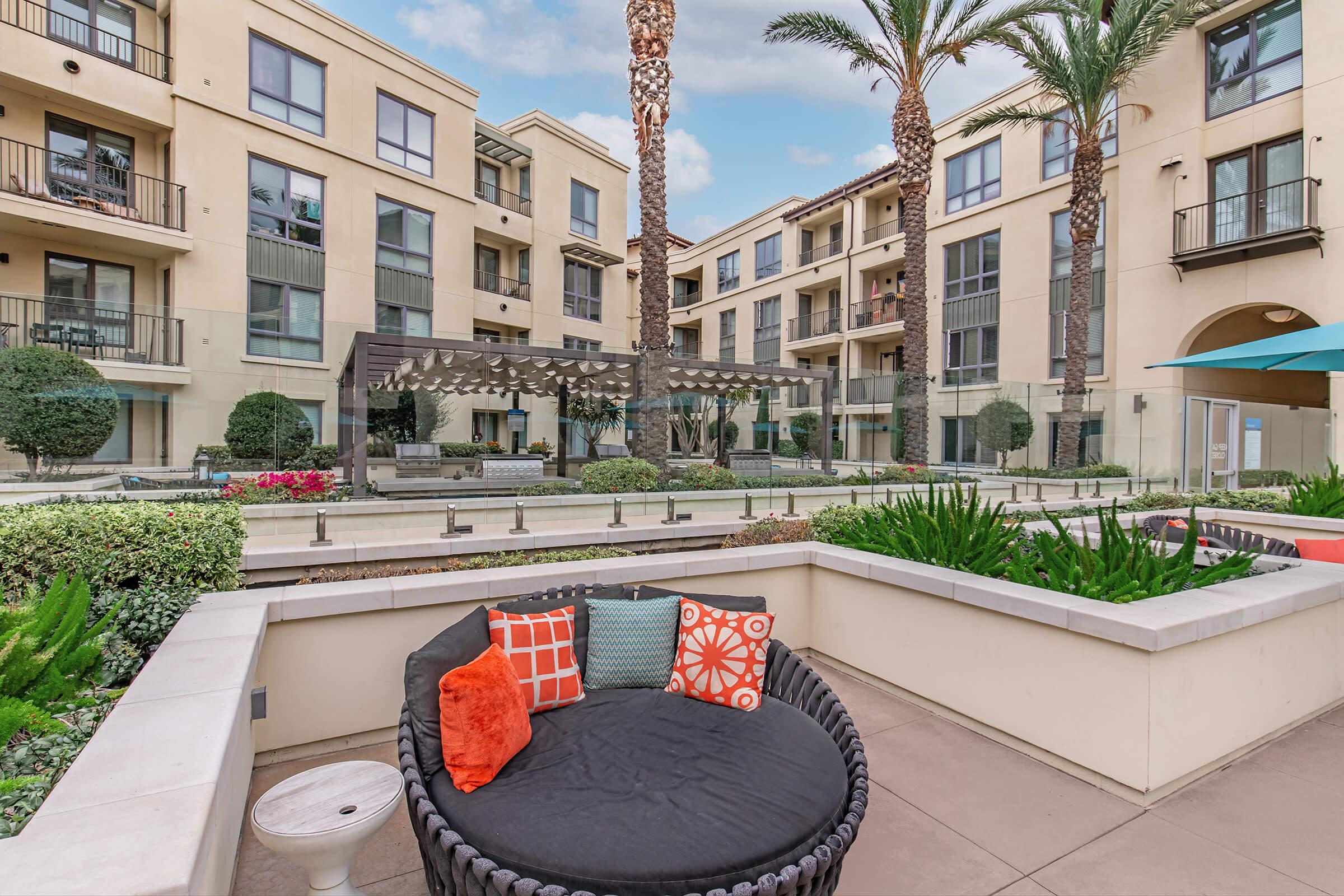 A modern outdoor lounge area featuring a circular black wicker sofa adorned with colorful cushions, surrounded by landscaped greenery, palm trees, and contemporary apartment buildings. The space includes a pergola and seating areas, creating a relaxed and inviting atmosphere.