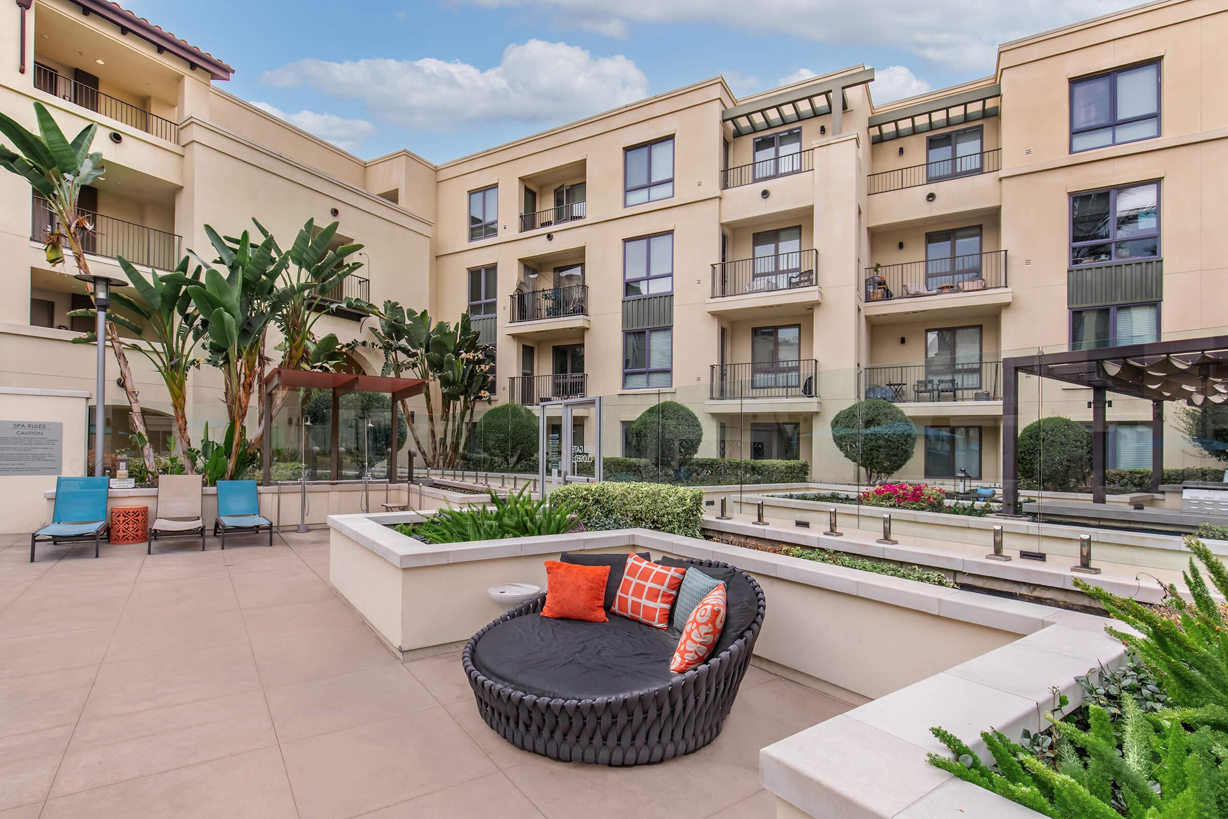 A modern apartment complex courtyard featuring landscaped gardens, seating areas, and an outdoor lounge chair with decorative pillows. Green shrubs and plants surround the space, with several balconies visible in the background, showcasing a contemporary architectural design.