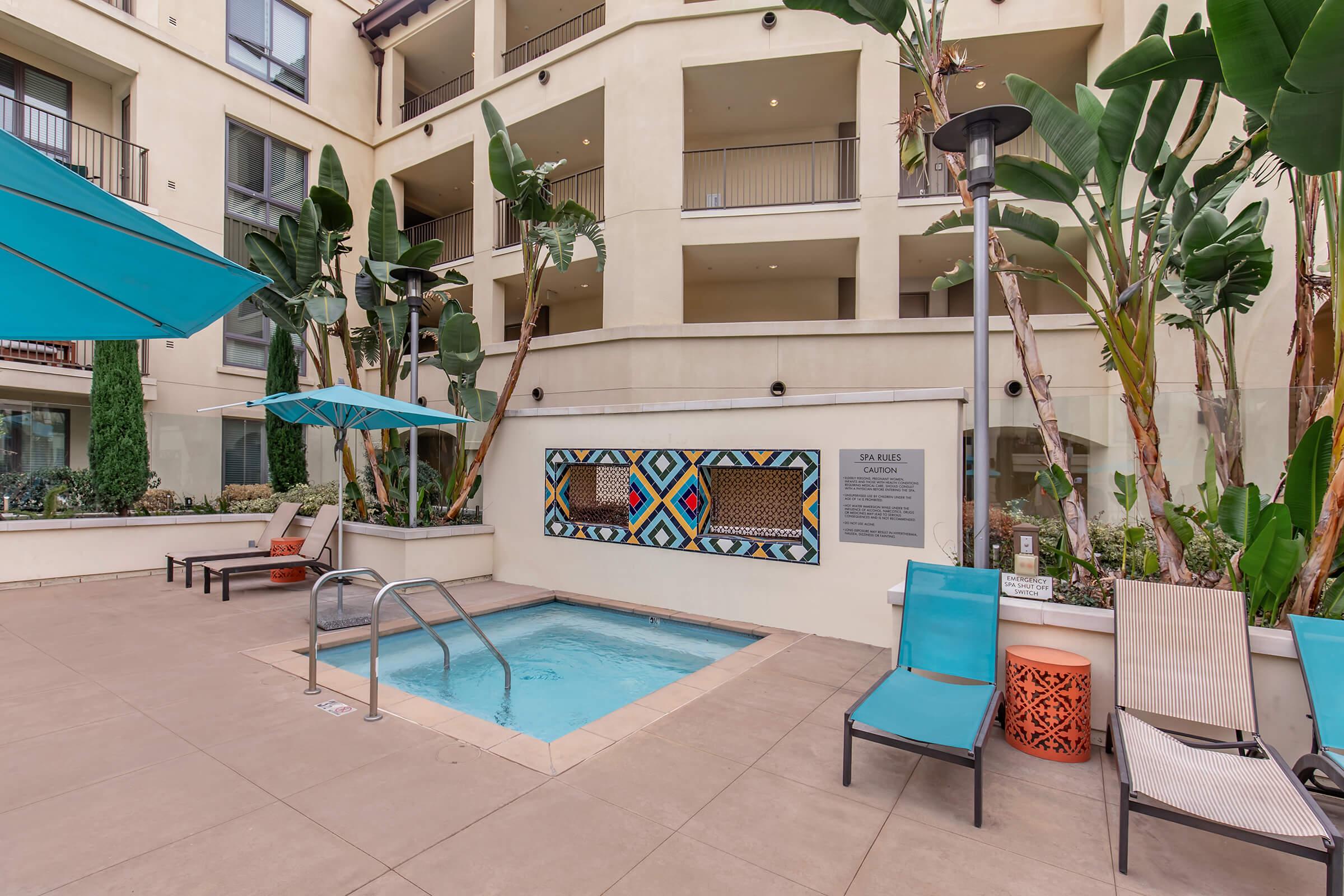A pool area in a modern residential complex, featuring a small rectangular pool, sun loungers, and vibrant tropical plants. Two teal umbrellas provide shade, and a colorful mural is displayed on the wall, adding artistic flair to the space. The setting is inviting and well-maintained.