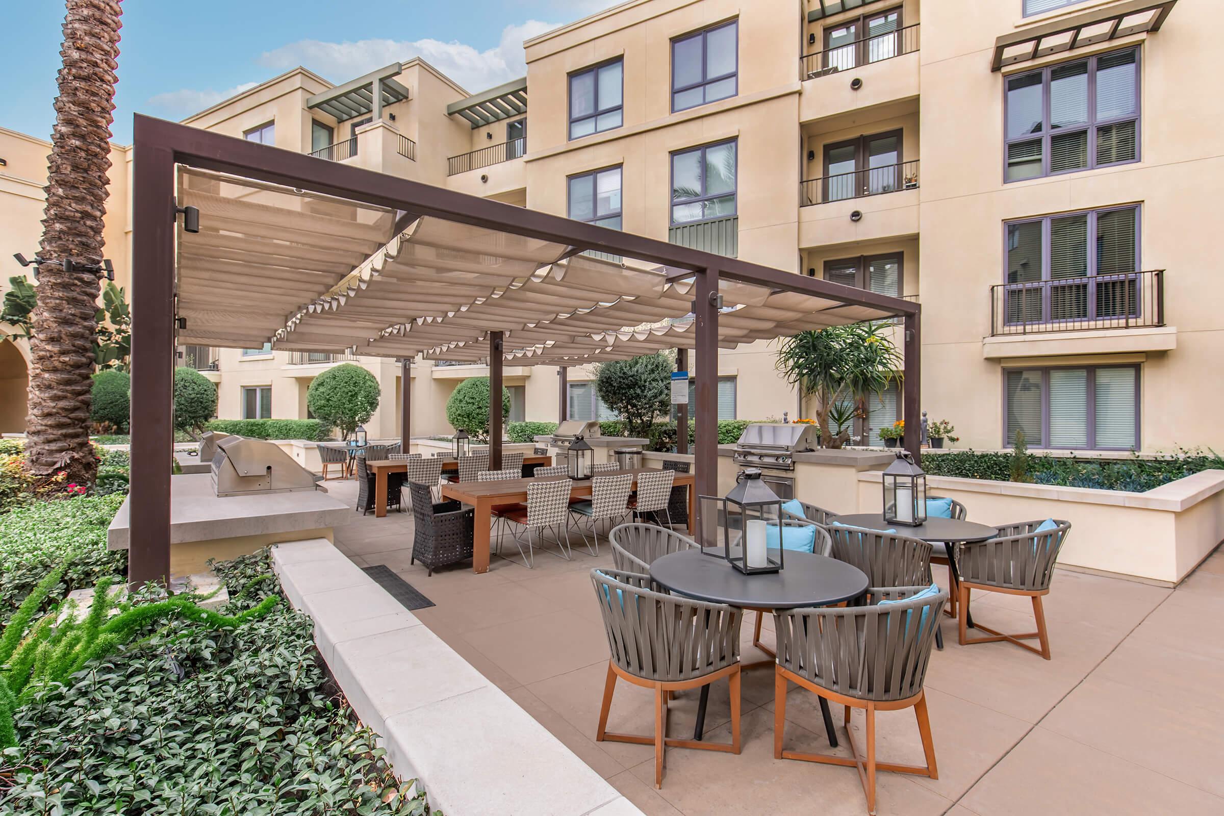 An outdoor seating area with round tables and chairs under a wooden pergola. Nearby, there are decorative lanterns and lush greenery. In the background, a modern apartment building with balconies and windows is visible. The setting is bright and inviting, suitable for social gatherings.