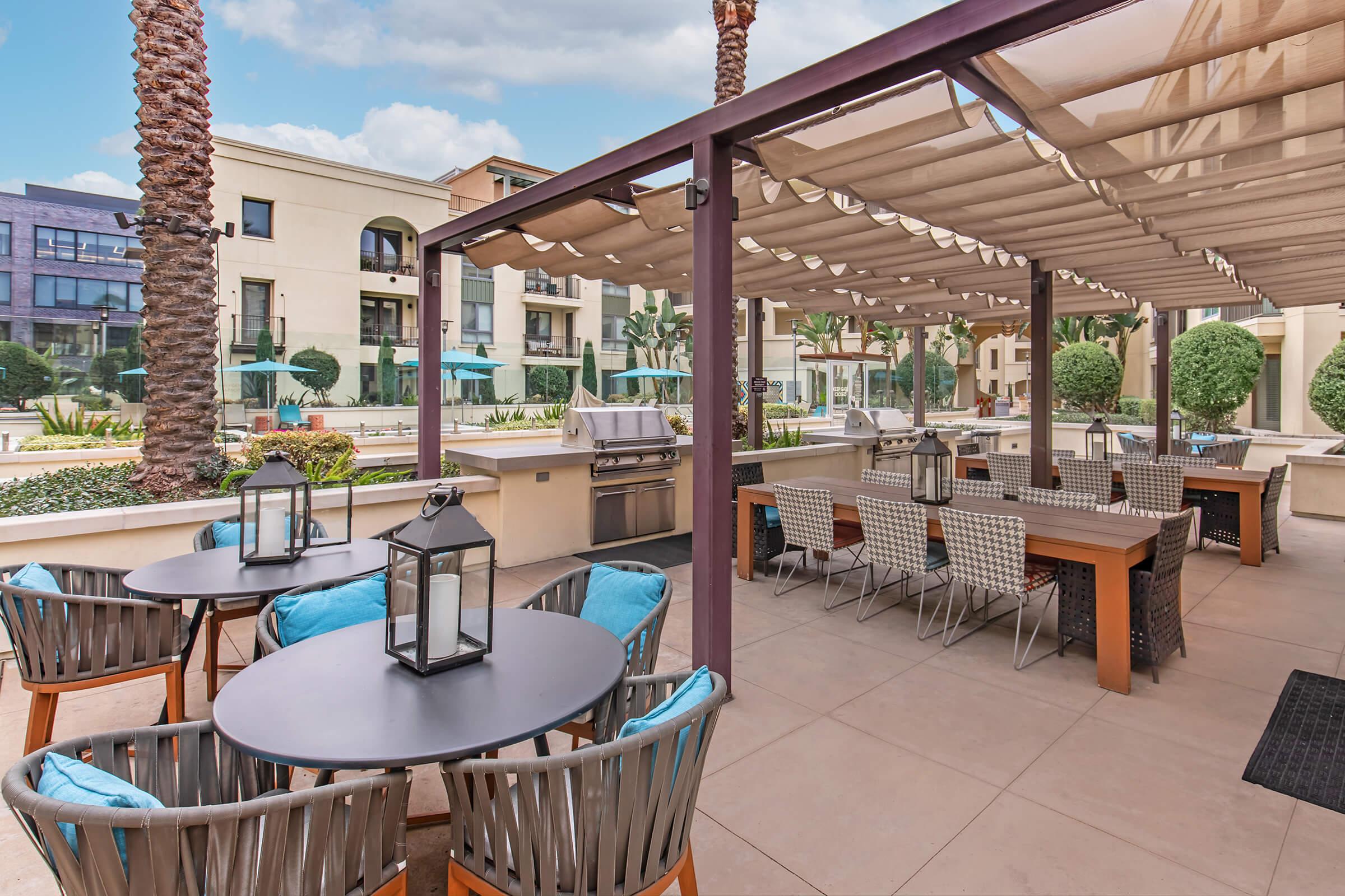 An outdoor dining area featuring stylish tables and chairs with blue cushions. There are decorative lanterns on the tables, a BBQ grill in the background, and lush landscaping with palm trees. The setting is bright and inviting, perfect for gatherings and meals in a relaxed atmosphere.