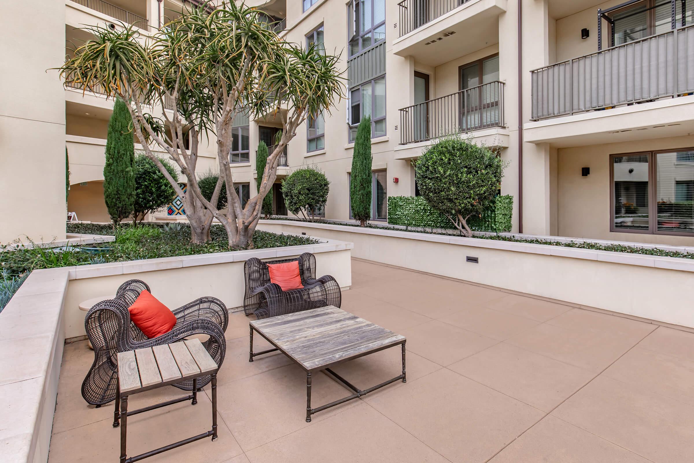 A cozy outdoor patio area with a small wooden table and two wicker chairs, one with red cushions. Surrounded by neatly trimmed shrubs and trees, the space is framed by modern apartment buildings, creating a comfortable and inviting atmosphere for relaxation.