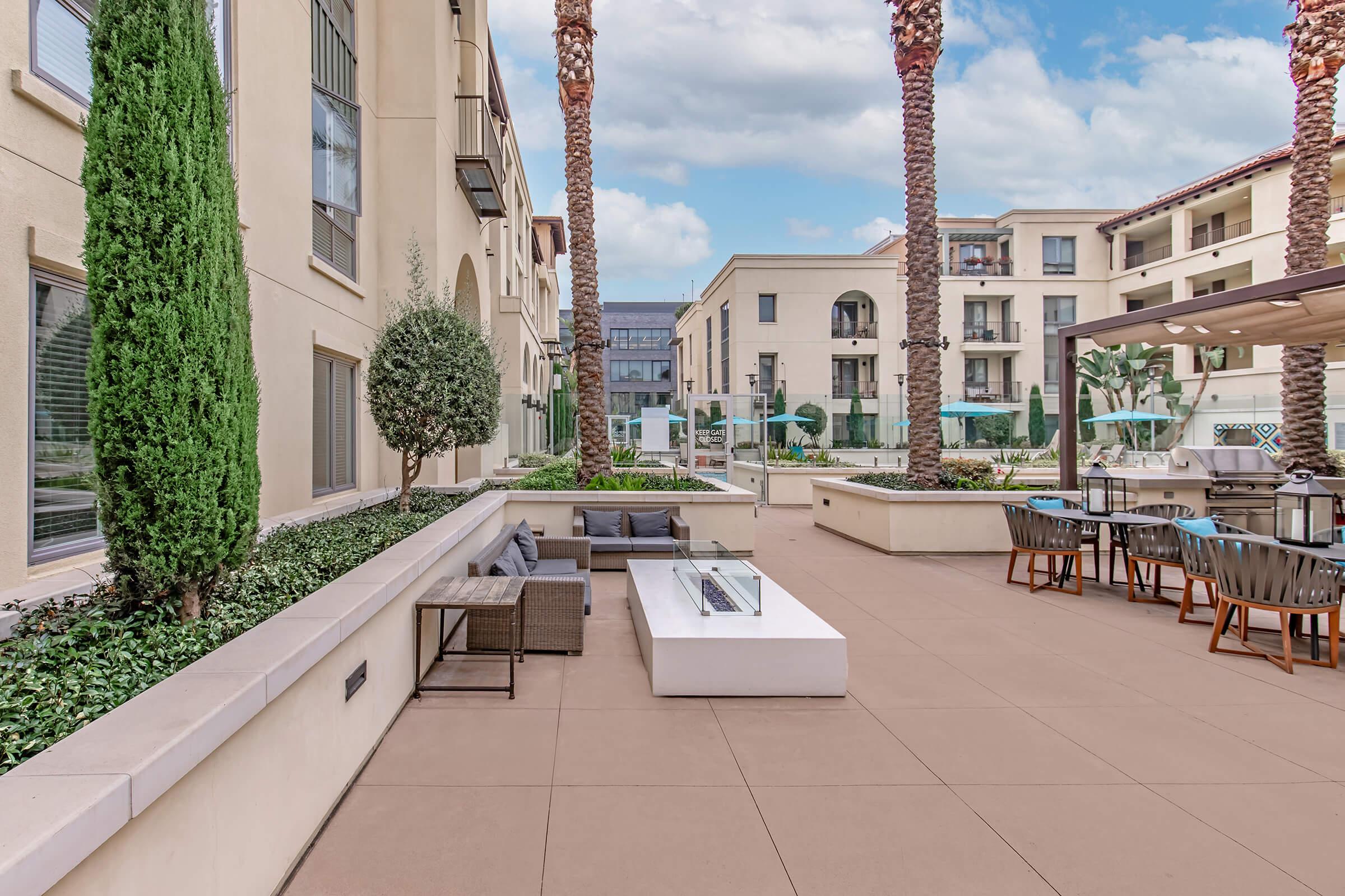 Outdoor courtyard area featuring palm trees, modern seating arrangements, a fire pit, and dining space. The surrounding buildings exhibit contemporary architecture, with outdoor amenities such as a pool in the background. The scene is set against a partly cloudy sky.