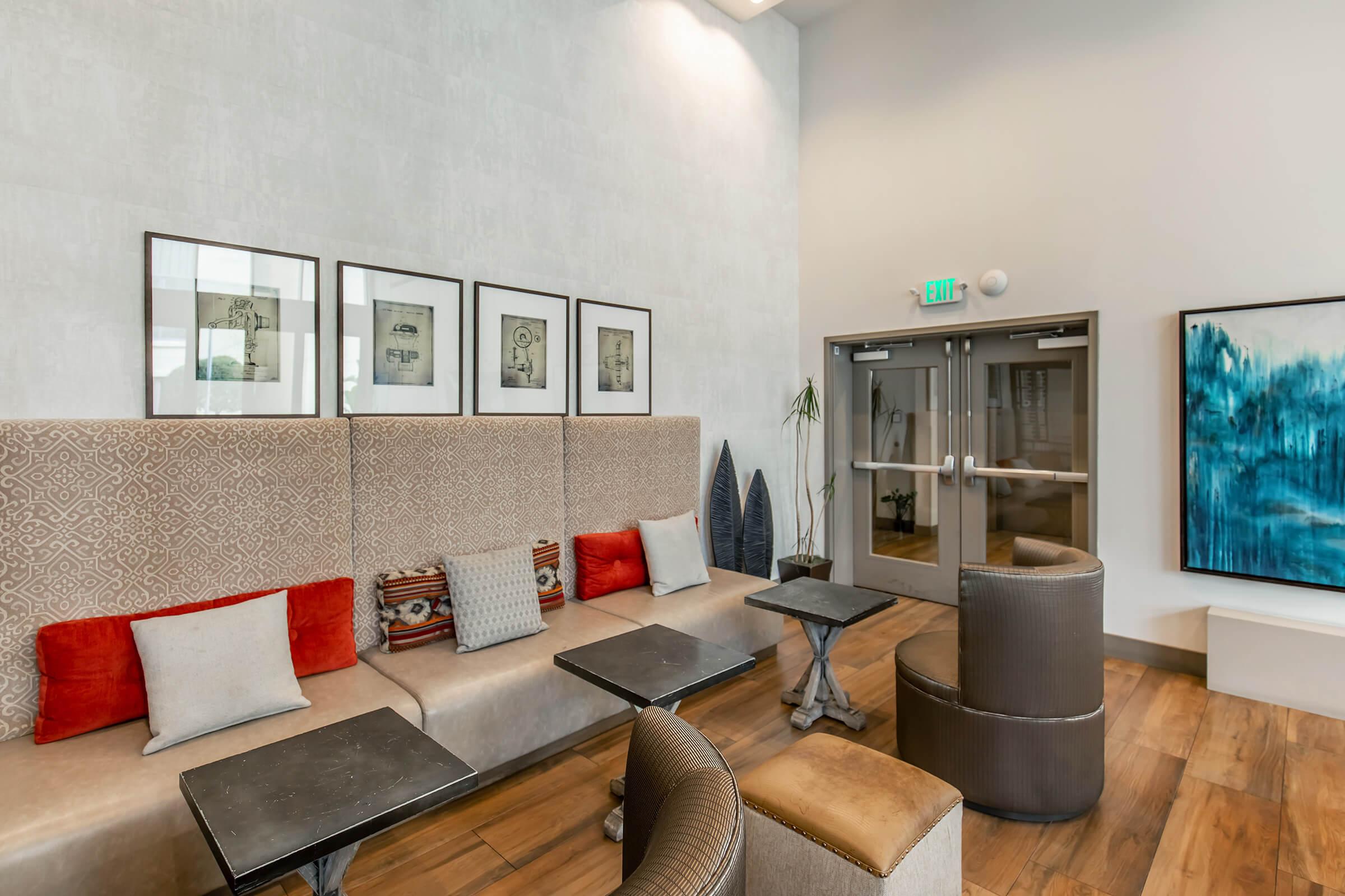 Modern lounge area featuring a comfortable seating arrangement with decorative cushions, small tables, and framed artwork on the walls. Large windows allow natural light, enhancing the inviting atmosphere. A painting in blue tones adds a focal point to the room, creating a contemporary and relaxed vibe.