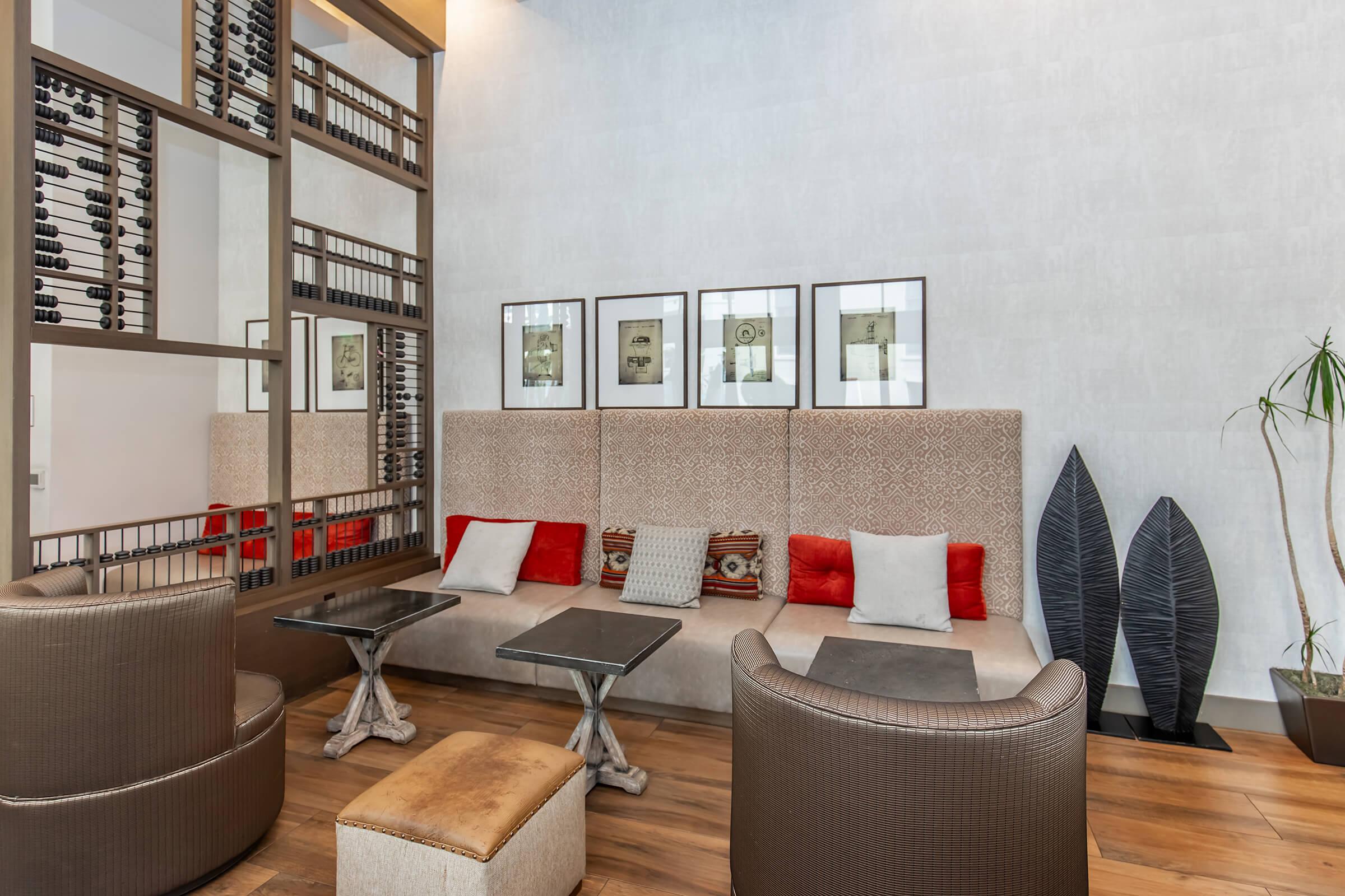 A modern lounge area featuring two upholstered chairs, a low table, and a stylish wall with framed artwork. The decor includes a mix of neutral and red tones, wooden flooring, and decorative plant elements. The space has a warm and inviting atmosphere, ideal for relaxation or social gatherings.