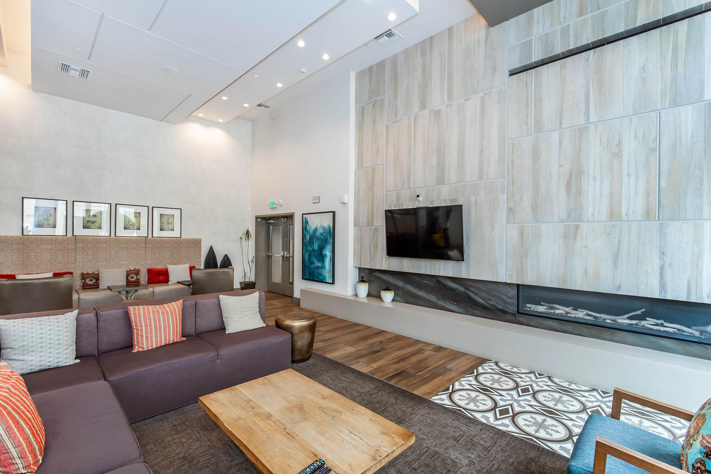 A modern, spacious living area featuring a cozy sectional sofa with decorative pillows, a wooden coffee table, and a contemporary fireplace. The walls have textured finishes and artwork. A large wall-mounted TV is visible, along with stylish light fixtures and patterned floor tiles.