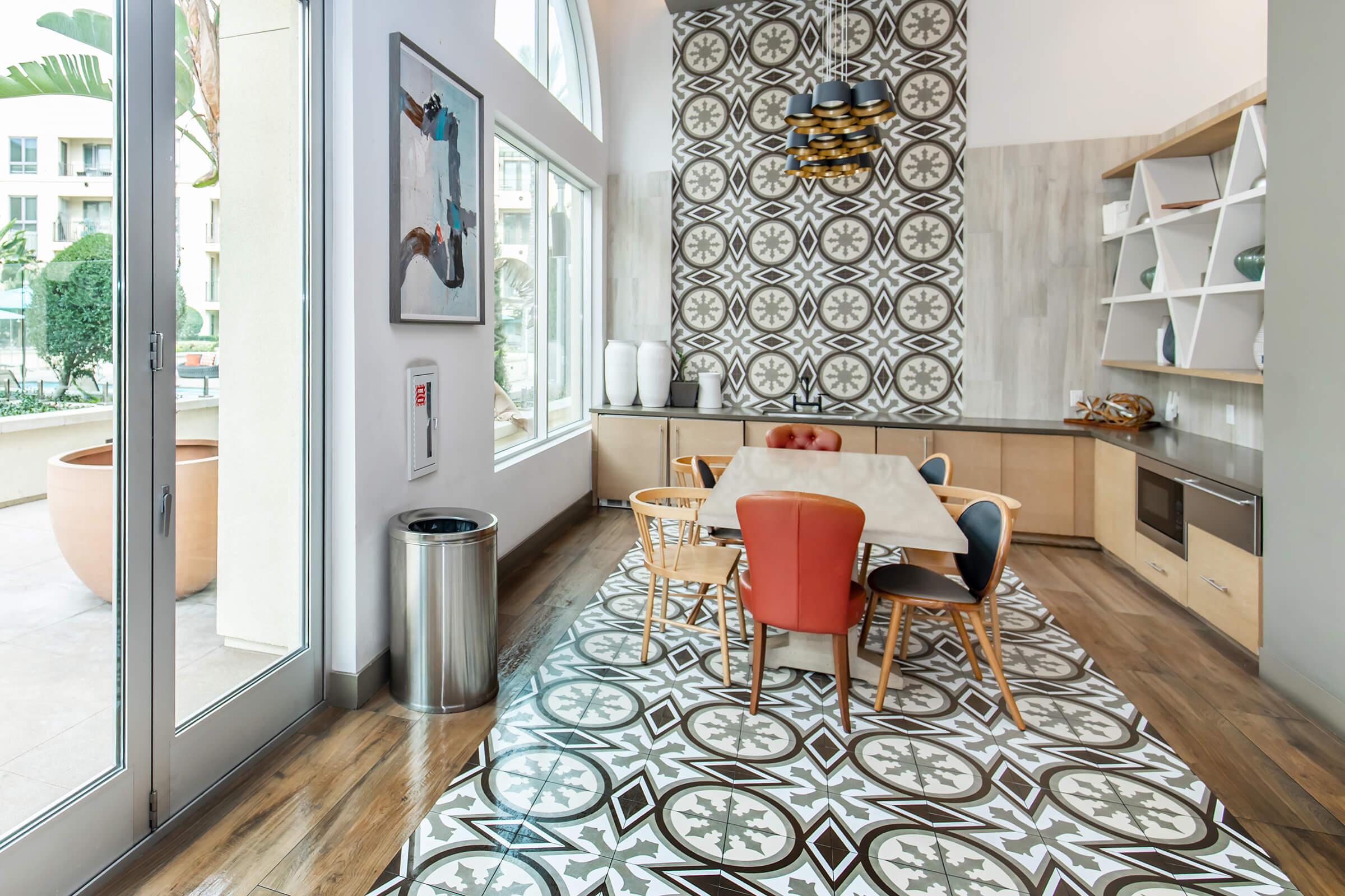 A modern dining area featuring a round table surrounded by colorful chairs, set against a patterned floor. Large windows allow natural light, and decorative elements include wall art and elegant shelving. A trash can and potted plants are visible, enhancing the contemporary aesthetic.