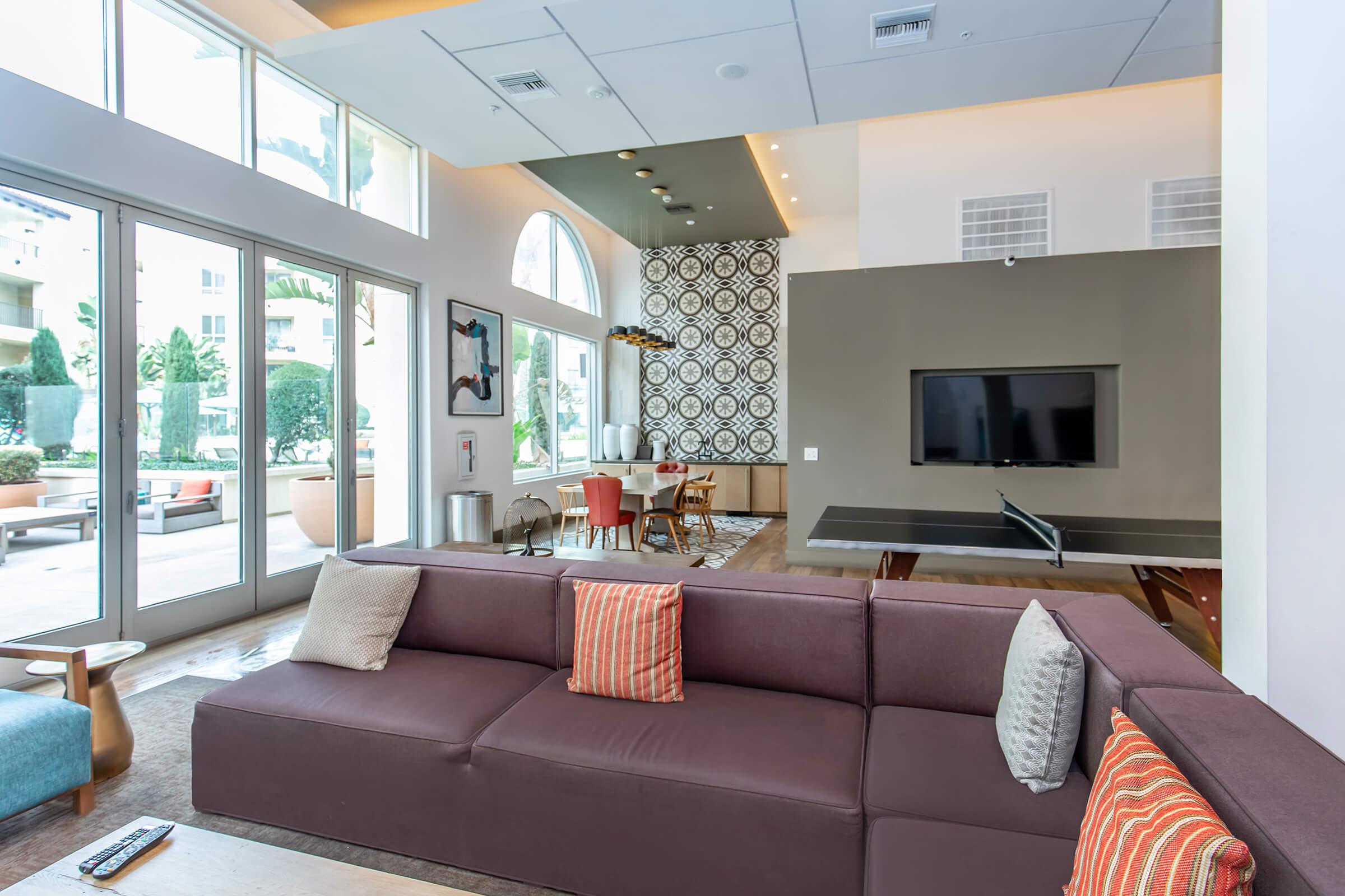 A modern living room featuring a large brown sectional sofa with colorful throw pillows. The room has large windows letting in natural light, a flat-screen TV mounted on the wall, and a dining area in the background. Decorative wall patterns add visual interest, and outdoor greenery is visible through the windows.