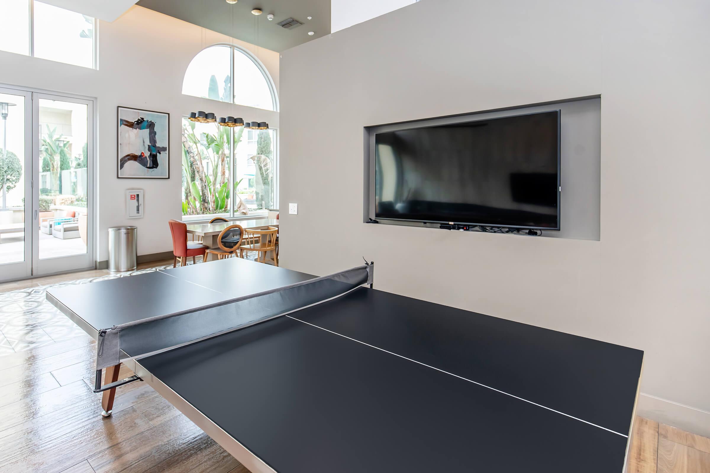 A modern indoor space featuring a ping pong table in the foreground, with a sleek television mounted on the wall. Large windows allow natural light to fill the room, showcasing greenery outside. Stylish decor elements can be seen on the walls and furniture arranged in the background.
