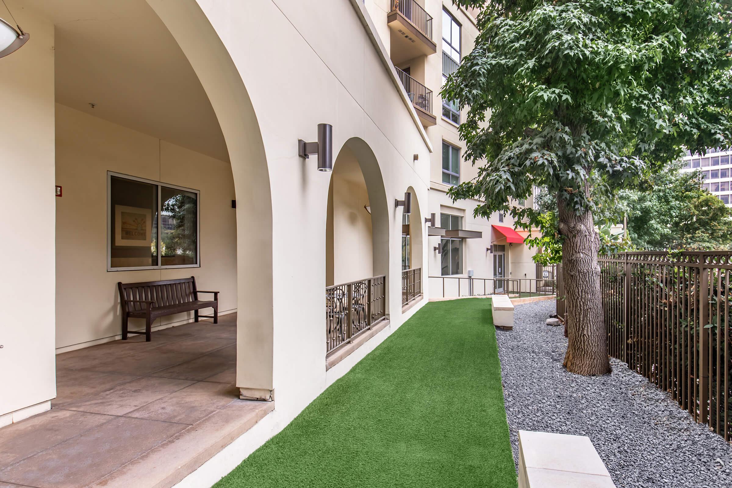 A view of a modern outdoor space featuring a pathway lined with synthetic grass, a bench under an archway, and landscaped areas with gravel and trees. The exterior of a building is visible, showcasing windows and a mixture of textures in the architecture.