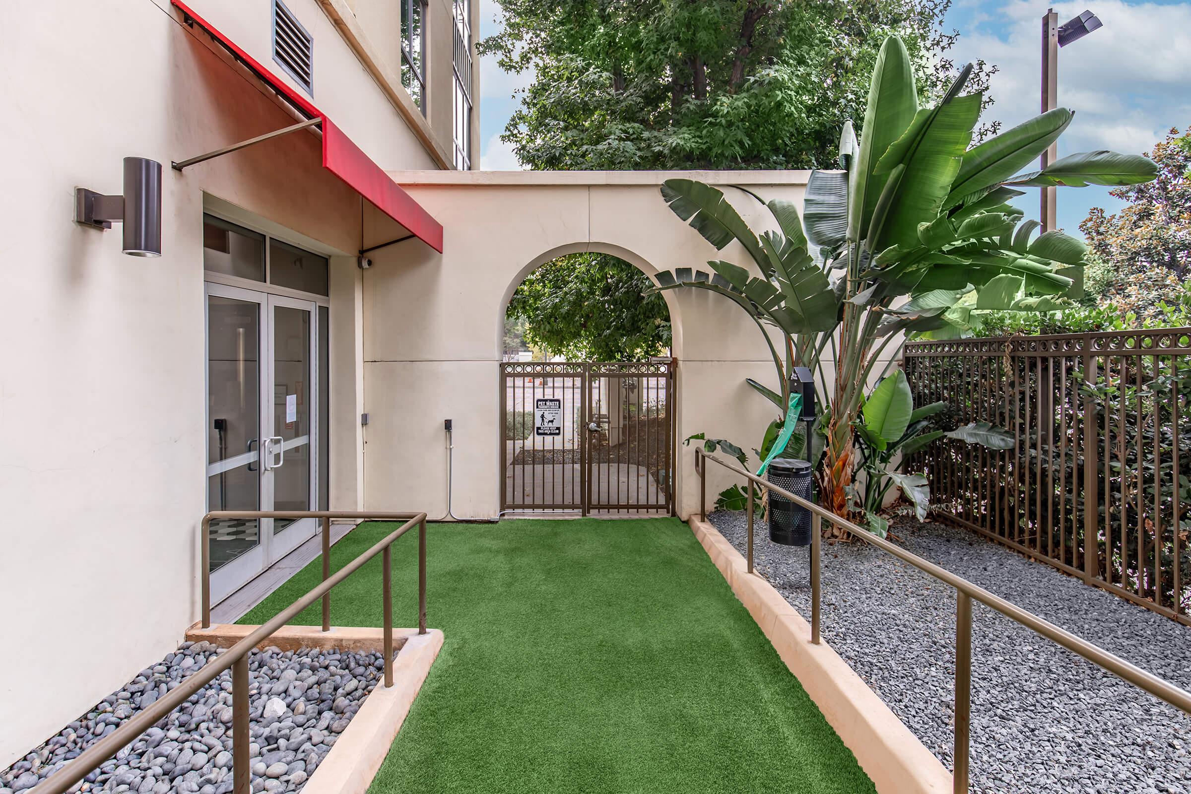 A landscaped entrance area with artificial grass and decorative gravel. There is a gated archway leading to a garden space, featuring tropical plants, and a building entrance with a red awning. The scene is bright and inviting, surrounded by greenery and modern architecture.