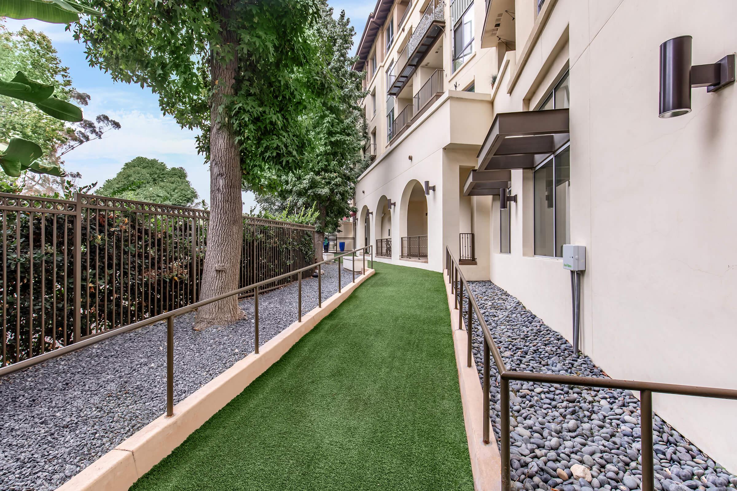 A narrow walkway lined with artificial grass and decorative gravel, flanked by a fence and large trees. The path leads alongside a modern building, characterized by its arches and windows. The scene is well-lit and appears tidy and inviting.