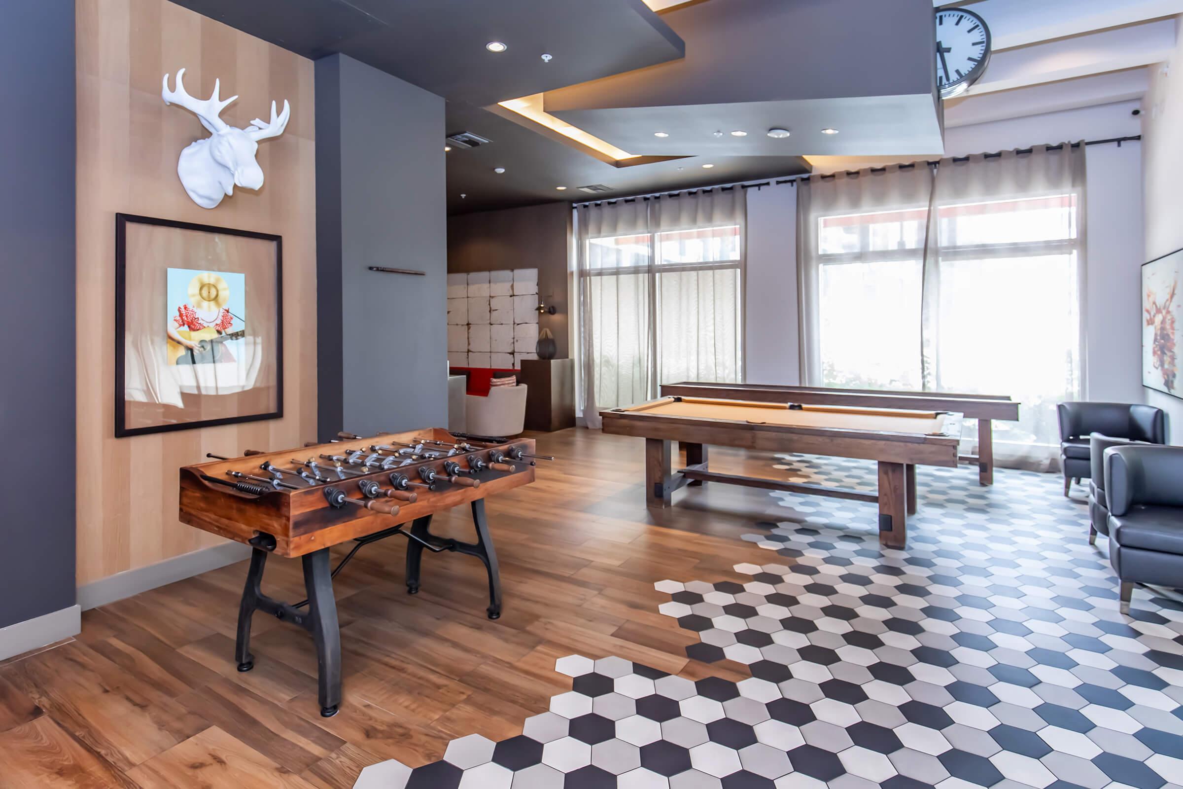 A modern game room featuring a foosball table and a pool table. The room has wood and hexagon-patterned tile flooring, with large windows allowing natural light. Decorative elements include a framed artwork and a mounted deer head, along with comfortable seating and stylish lighting fixtures.