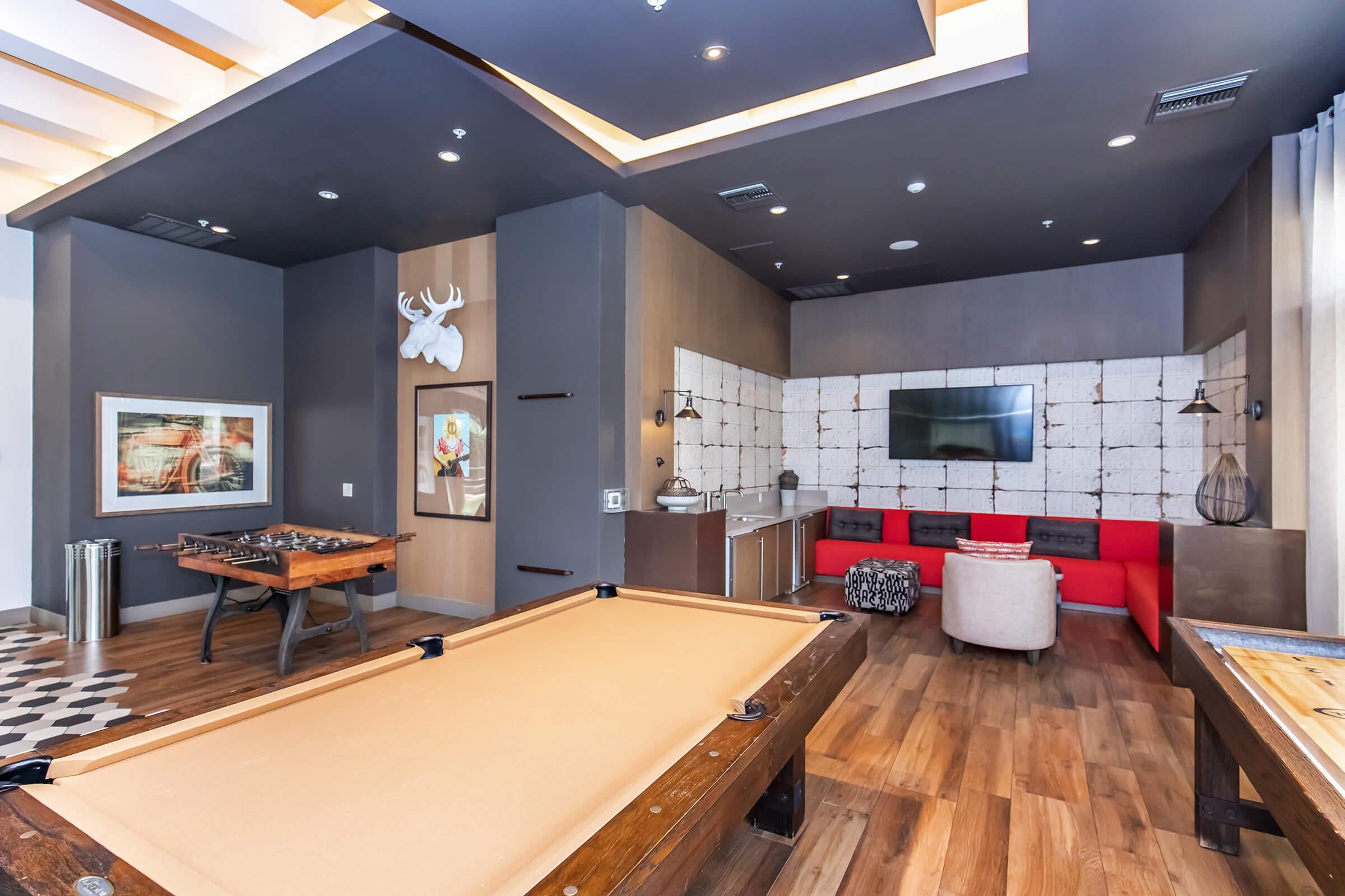 A stylish recreation room featuring a billiards table, foosball table, and a cozy seating area with red couches. The walls are decorated with modern art and a mounted deer head, while a wall-mounted TV adds to the entertainment options. The flooring is made of rich hardwood.