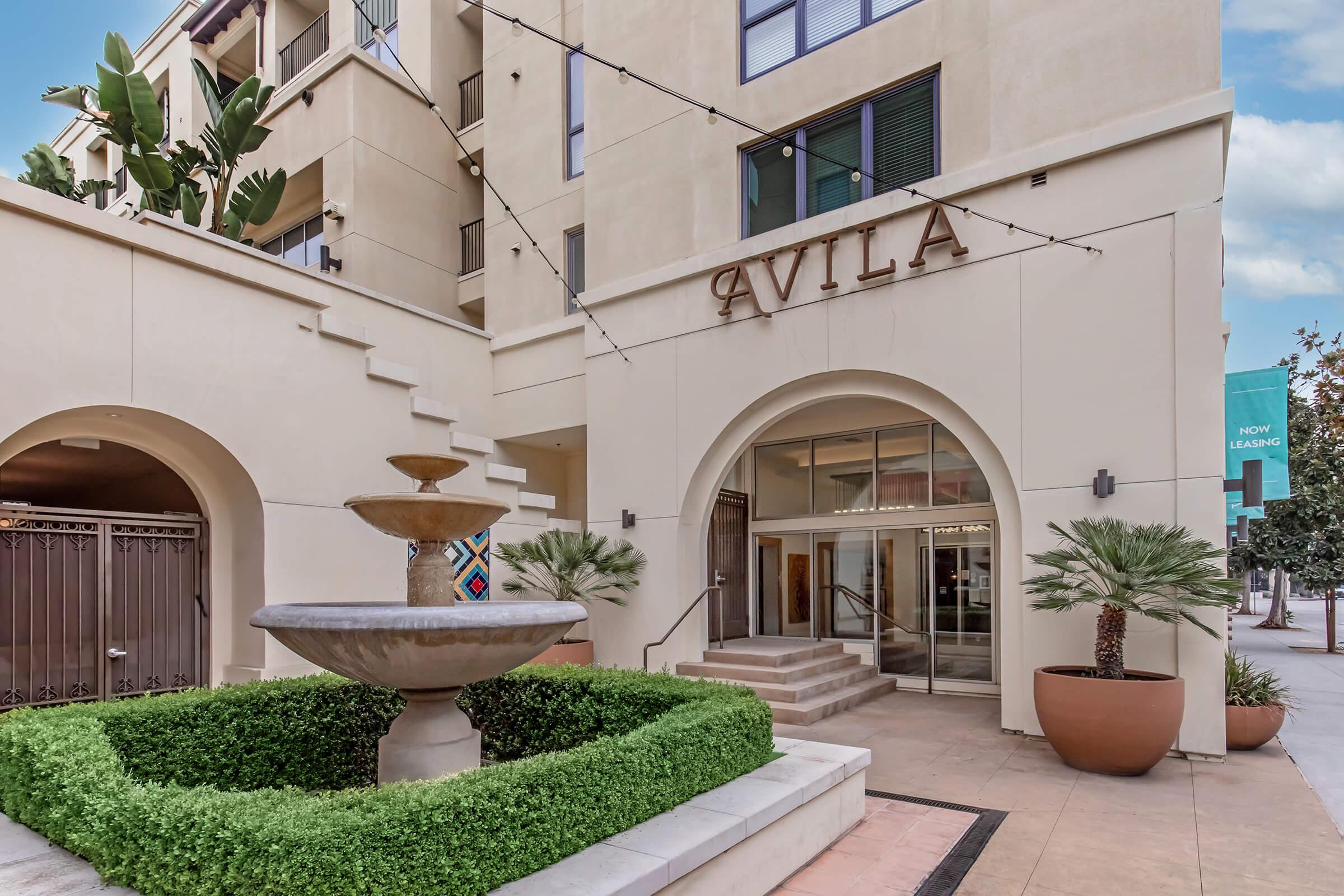 Entrance of a modern building named "Avila," featuring an elegant fountain surrounded by lush greenery and decorative plants. The steps lead to a welcoming lobby, with string lights overhead and a leasing banner visible. The architecture is contemporary with warm colors and inviting design elements.