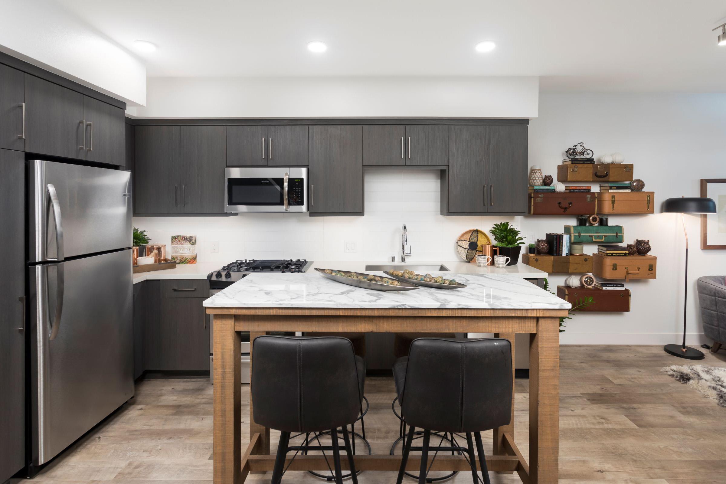 Modern kitchen featuring dark cabinetry, stainless steel appliances, and a marble countertop island with two black bar stools. The space is bright with recessed lighting and decorated with plants and wooden boxes for storage. A cozy living area is visible in the background.