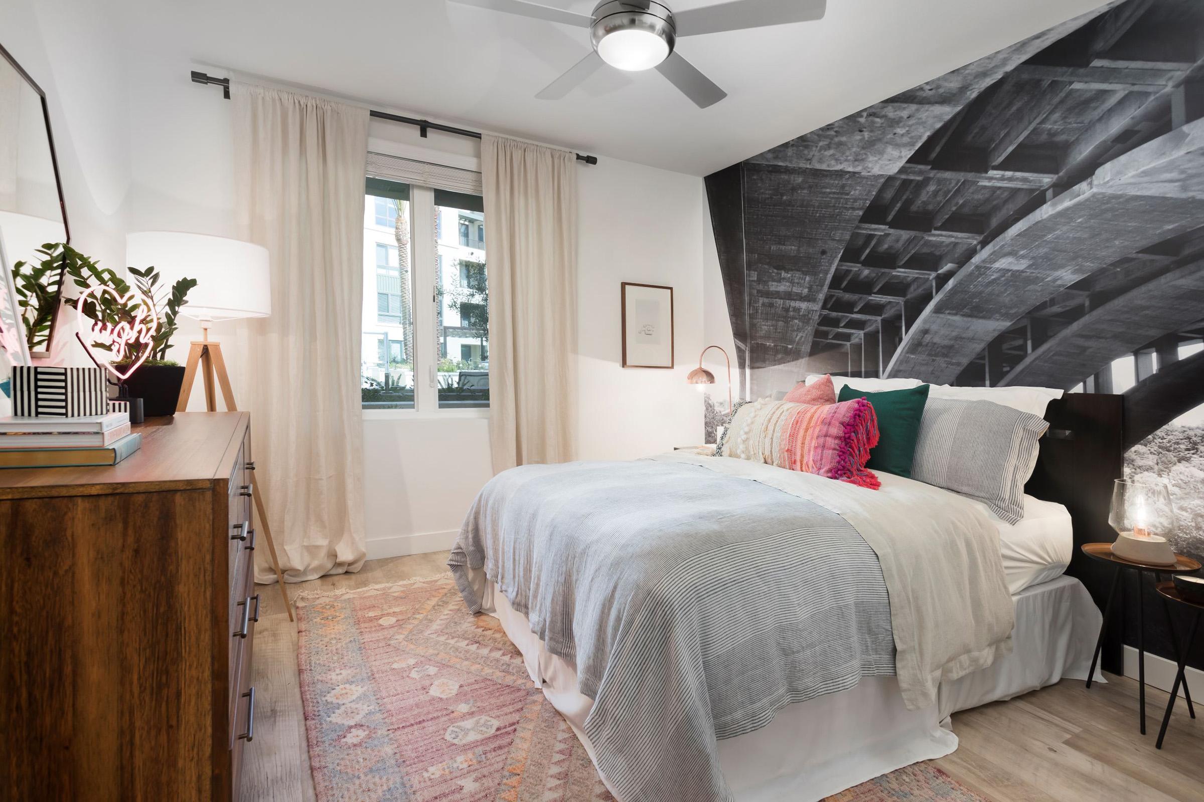 Cozy bedroom featuring a comfortable bed with layered bedding and colorful pillows. A stylish wooden dresser is to one side, while a lamp and decorative plants add charm. The wall behind the bed displays a dramatic black-and-white mural, and soft natural light filters through the curtains.
