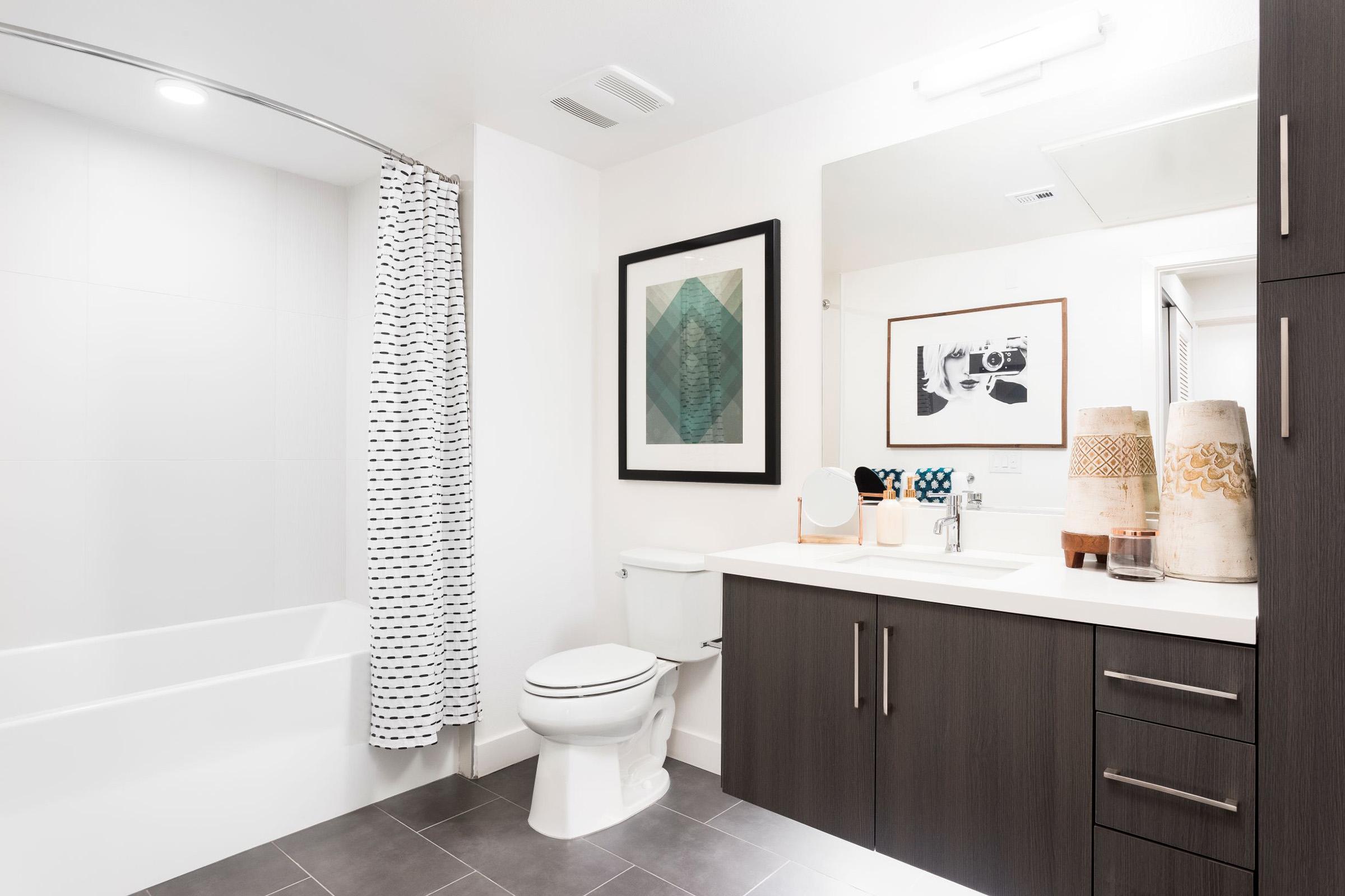 A modern bathroom featuring a white tub with a shower curtain, a sleek vanity with a sink, a large mirror, and decorative items on the countertop. The walls are mostly white, with dark cabinetry and a geometric art piece hanging nearby, complemented by neutral-toned tiles on the floor.