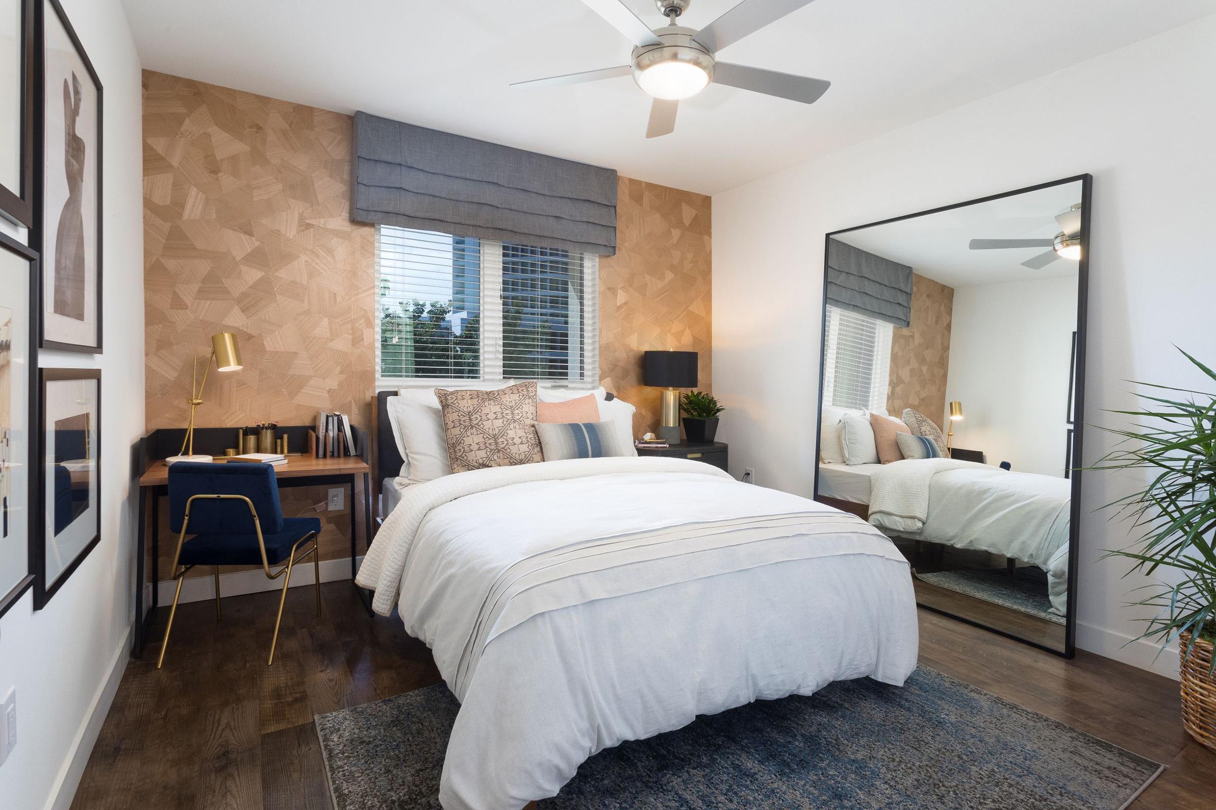 A modern bedroom featuring a plush bed with white linens, decorative pillows, and a stylish patterned wall. A large mirror reflects the space, while a small desk with a blue chair and brass lamp is positioned nearby. Natural light streams in through the windows, complemented by indoor plants and minimalist decor.