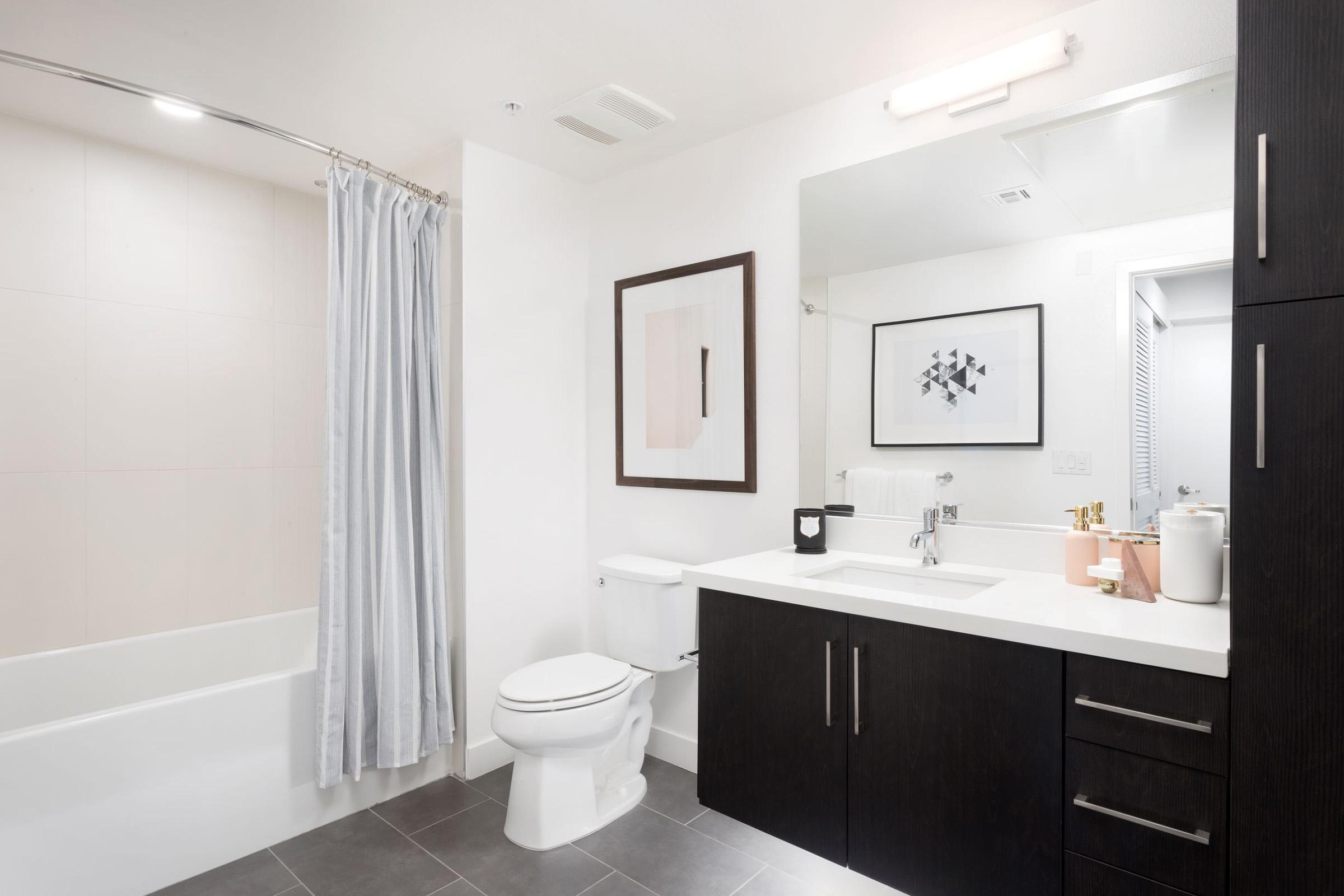 Modern bathroom featuring a bathtub with a light-colored shower curtain, a sleek vanity with dark wood cabinetry, a large mirror, and minimalistic decor, including framed artwork and a small vase. The space is well-lit with neutral tones and contemporary design elements.