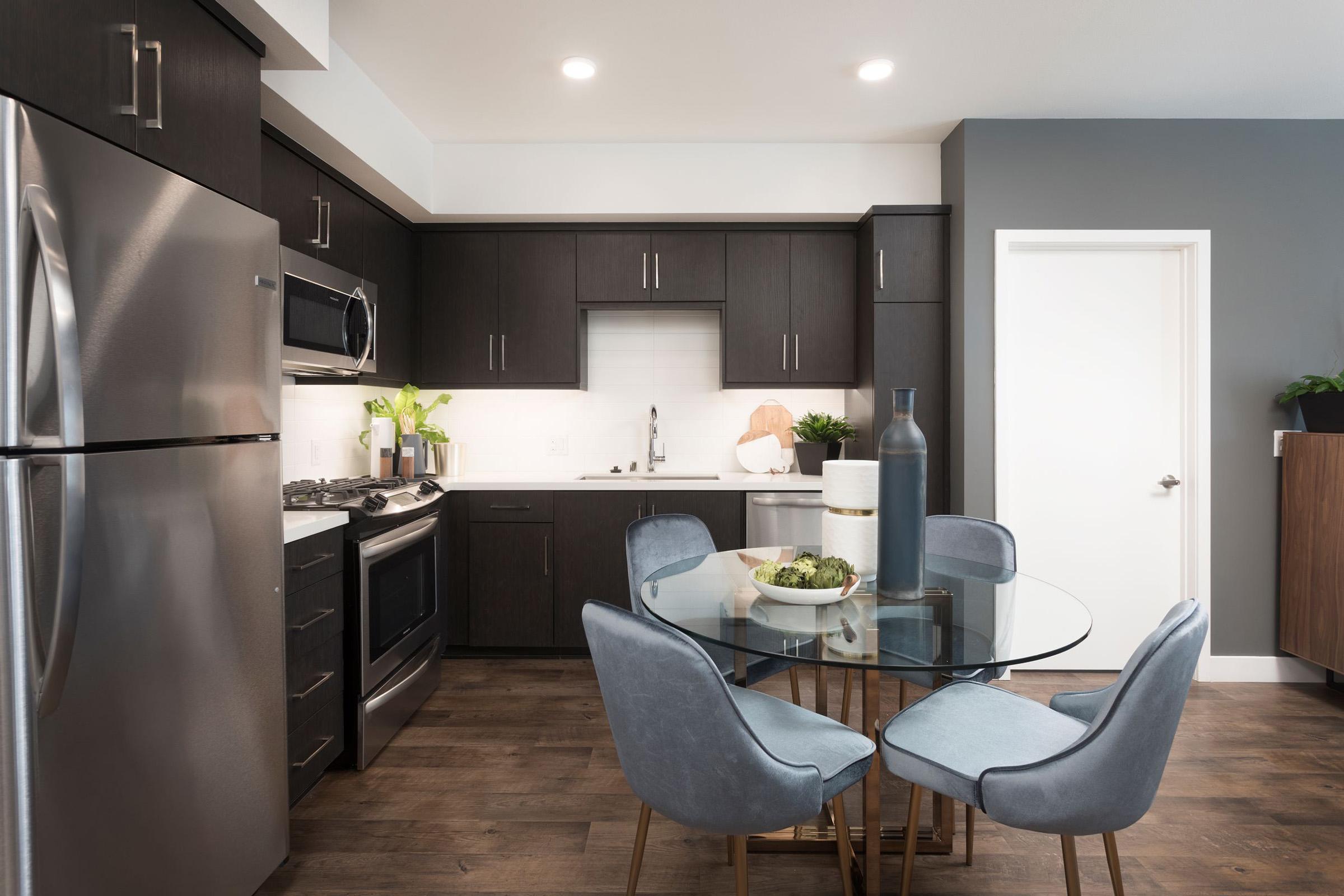 Modern kitchen featuring dark cabinetry and stainless steel appliances. A round glass dining table surrounded by four upholstered chairs is set in the center, with a decorative bowl of fruit. Natural light illuminates the space, accentuating the sleek design. A door leads to another room.